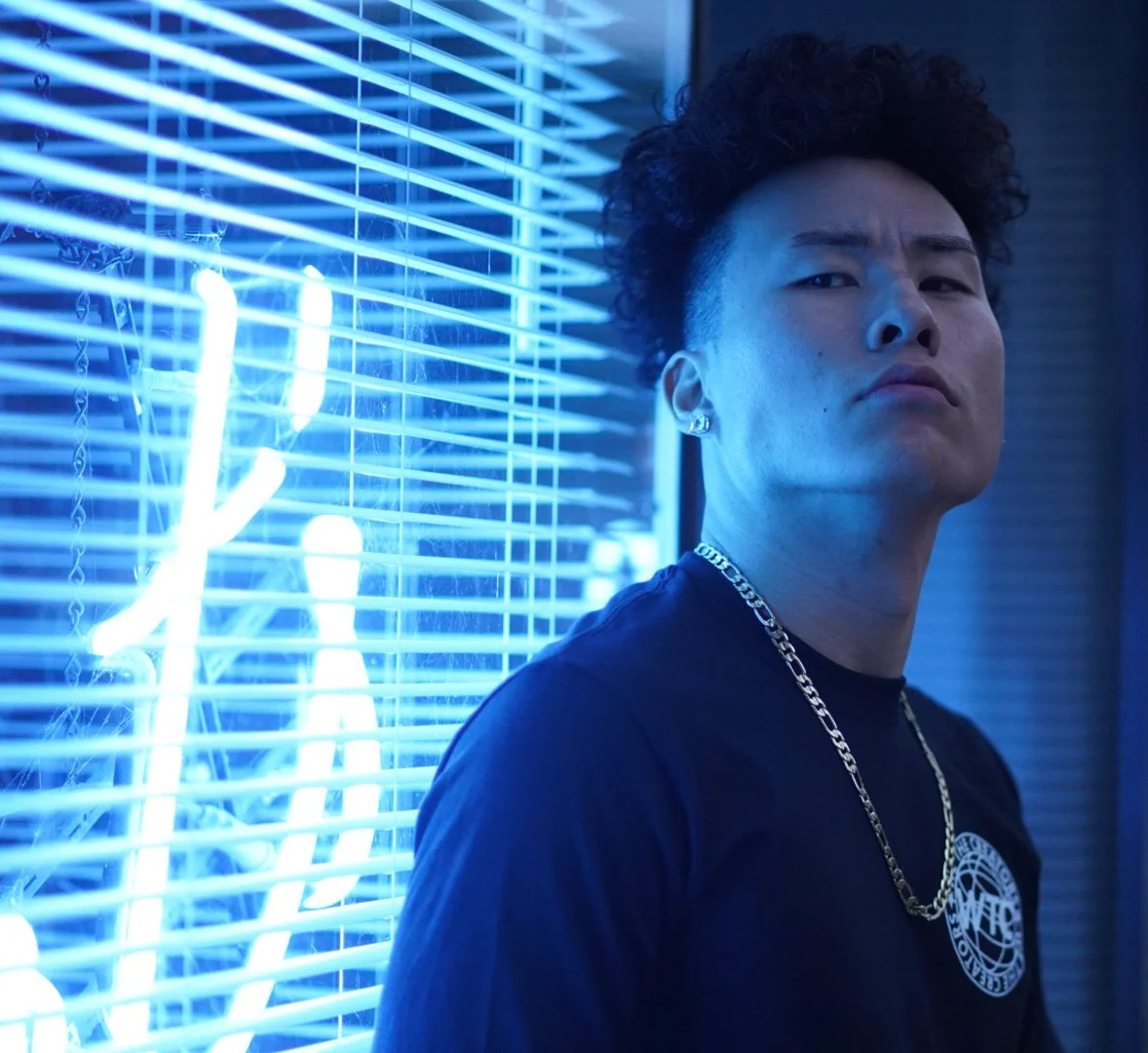 Young man with curly hair and earrings looking serious, wearing a chain necklace and a black shirt, standing by a blue neon light sign.