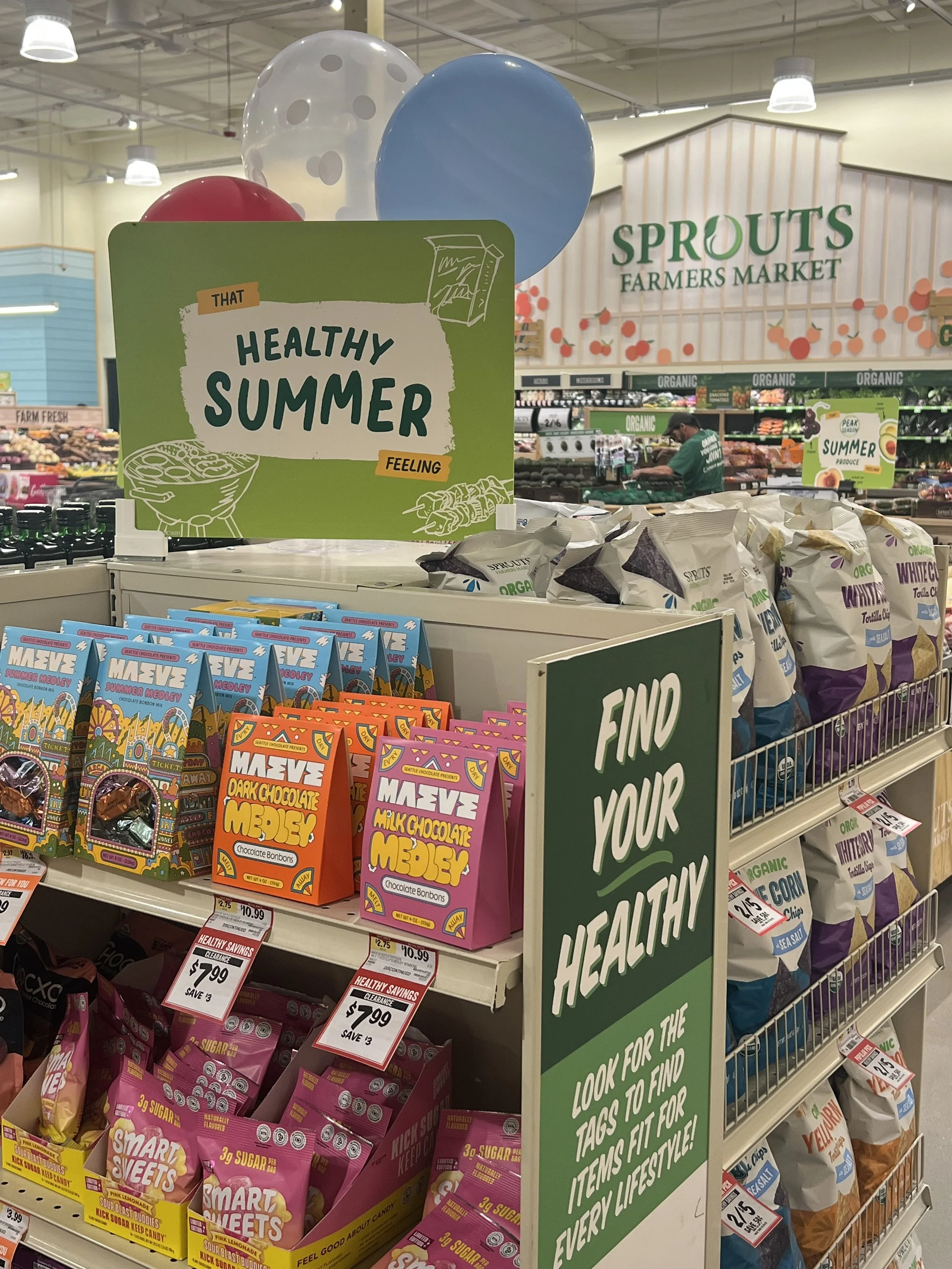 Shelf with colorful snack packages and signs promoting healthy summer eating at Sprouts Farmers Market.