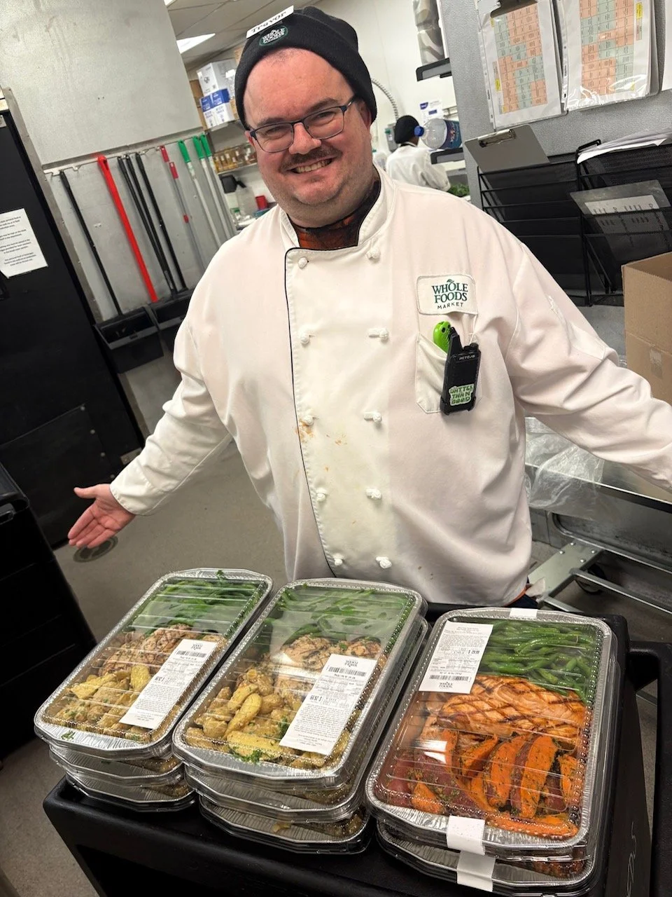 Trevor, smiling, with arms open, in a black hat and a white Whole Foods chef's coat.  In front of him, a selection of prepared foods in packaging.