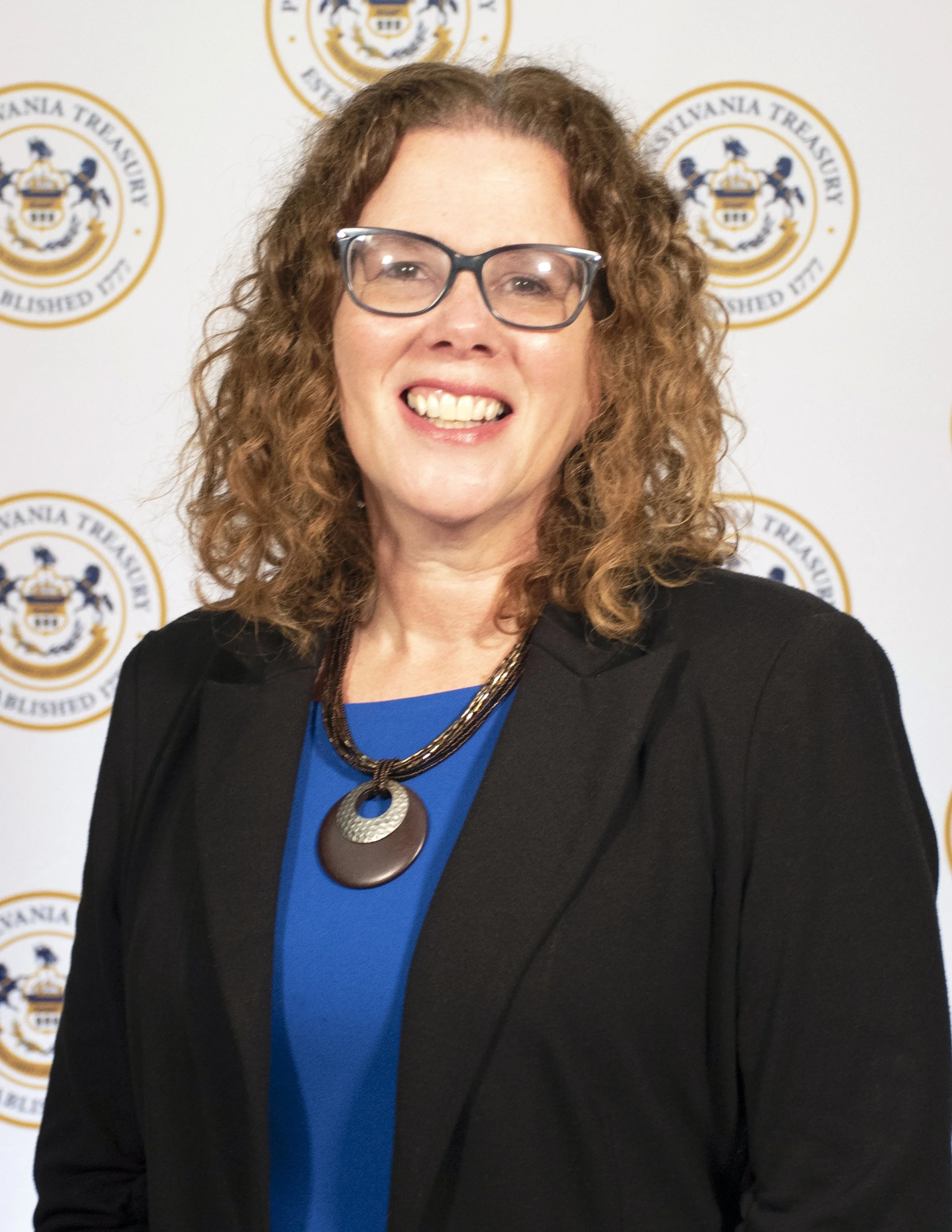 Photograph of Diana Fishlock, wearing a black blazer, a blue shirt, a bronze necklace, and dark-framed glasses.   She is looking at the camera and smiling, standing in front of a banner with the logo of the Pennsylvania treasury