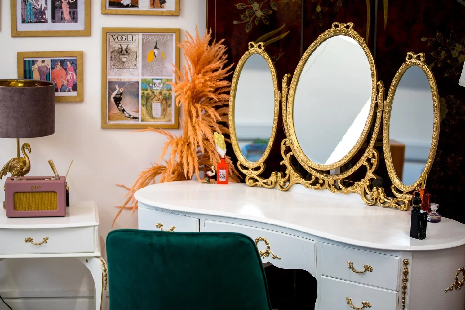 A colourful vanity cabinet with gold mirror, green chair, gold-framed artwork, and room divider