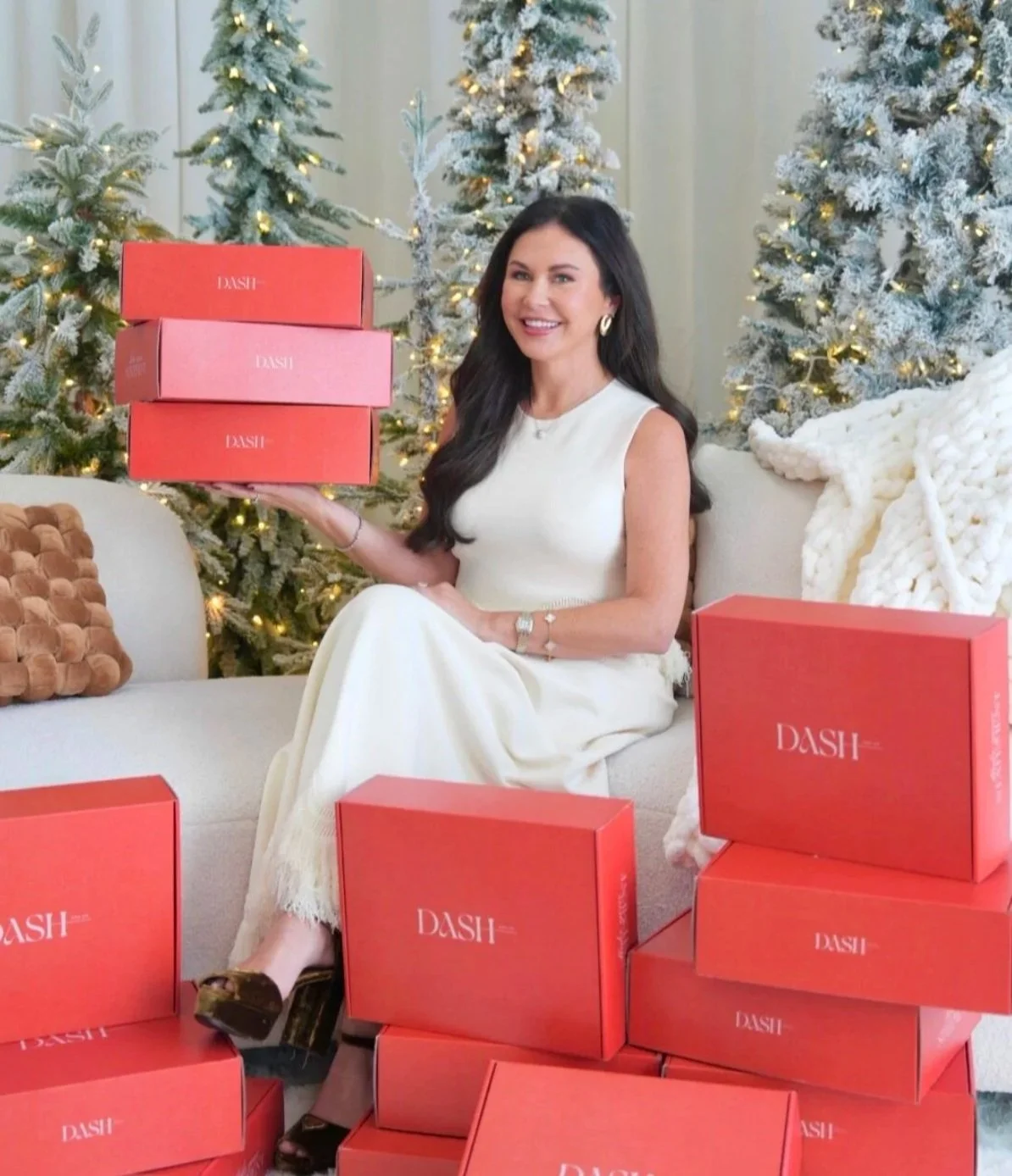 A woman with long dark hair sitting on a couch, holding a stack of three pink and red boxes labeled DASH. There are more DASH boxes around her on the floor. In the background, there are decorated Christmas trees with lights and a cozy blanket, indicating a festive setting.