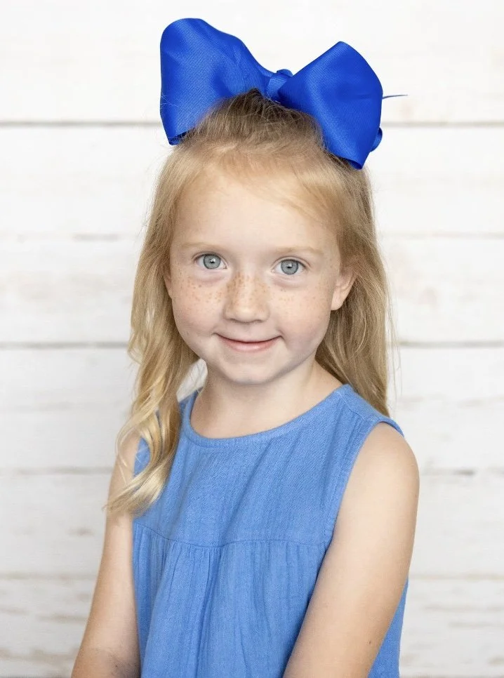 A young girl with blonde hair, blue eyes, and freckles, wearing a sleeveless blue dress and a large blue bow in her hair, standing in front of a white wooden background.