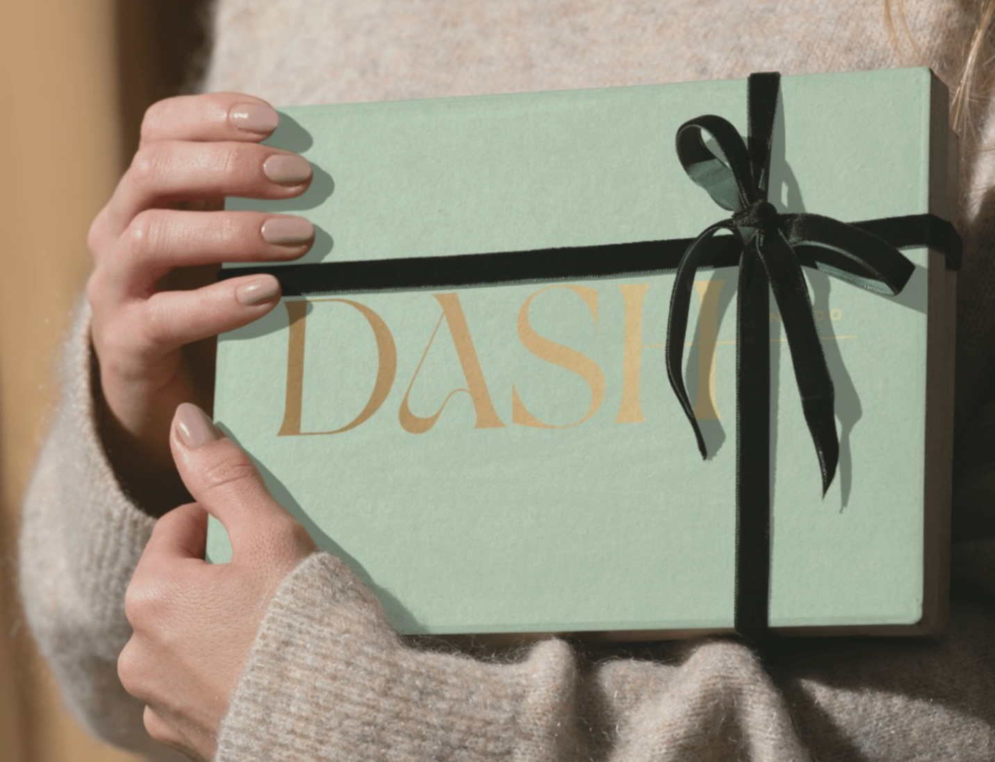 Person holding a pastel green gift box with the word "DASH" written in gold letters, wrapped with a black ribbon tied in a bow.