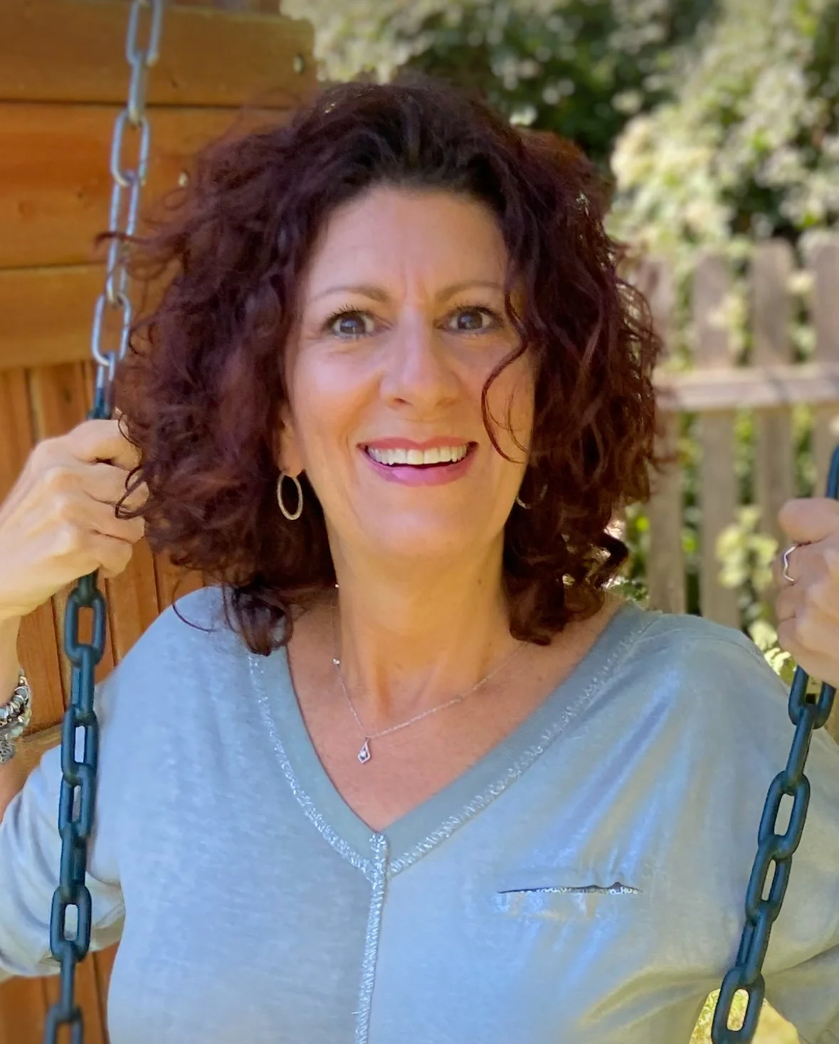A woman with curly brown hair smiling while sitting on a swing outdoors, holding the swing's chains with both hands, with a wooden fence and trees in the background.