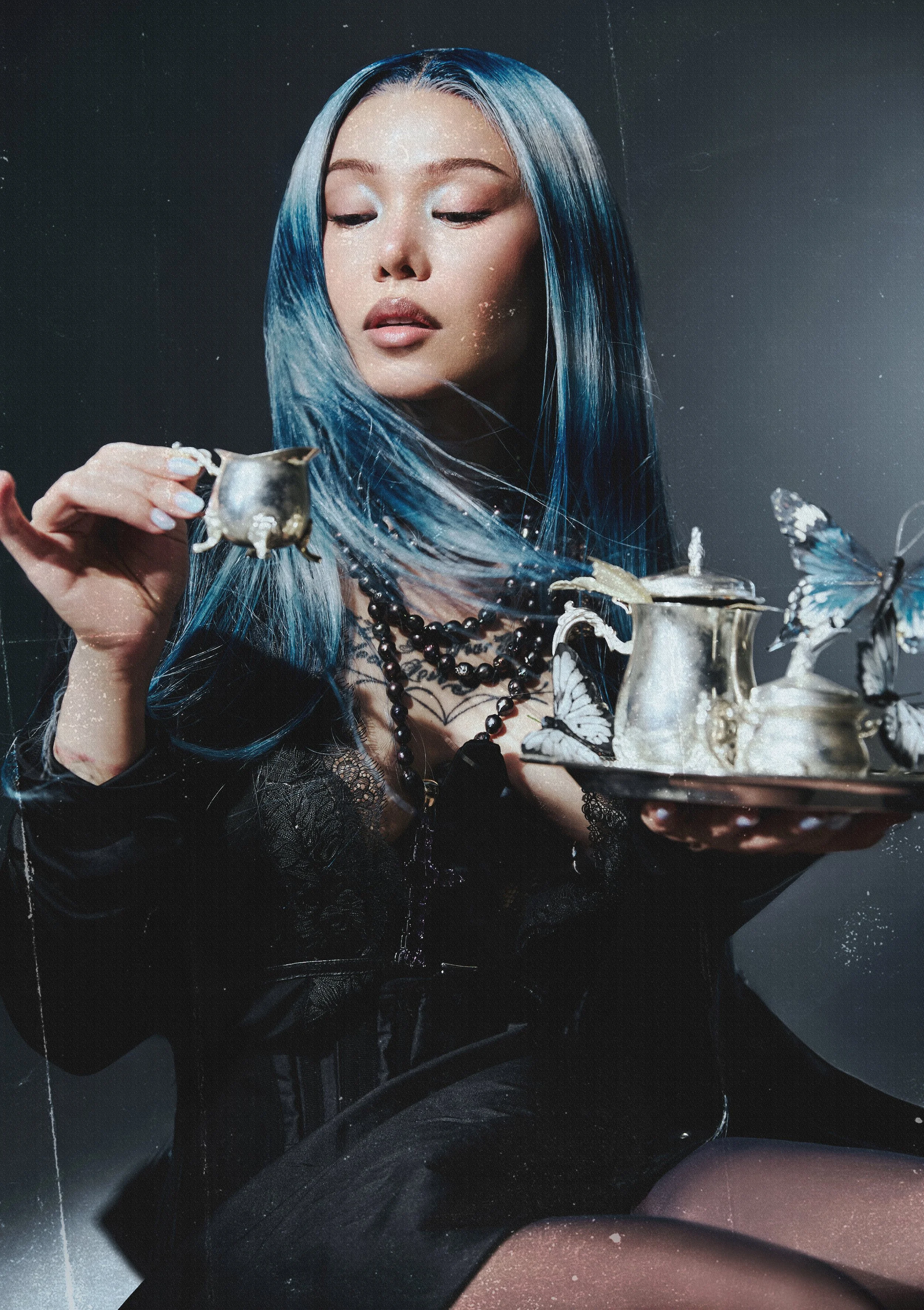 A woman with long, blue-tinted hair looking down at a silver tray with butterfly decorations and a silver pitcher, wearing a black lace dress and layered necklaces, against a dark background.