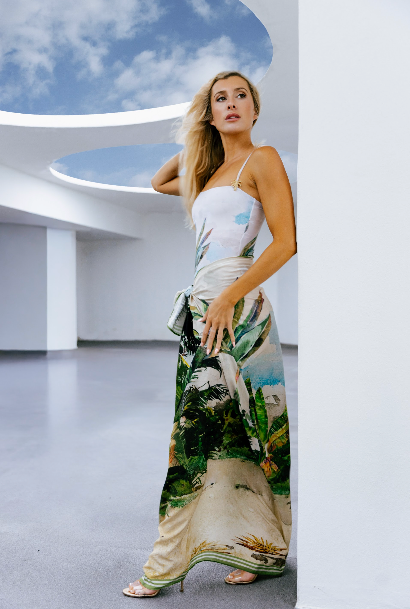 A woman in a long, colorful, tropical print dress standing indoors against a white wall with a circular skylight showing a blue sky with clouds.