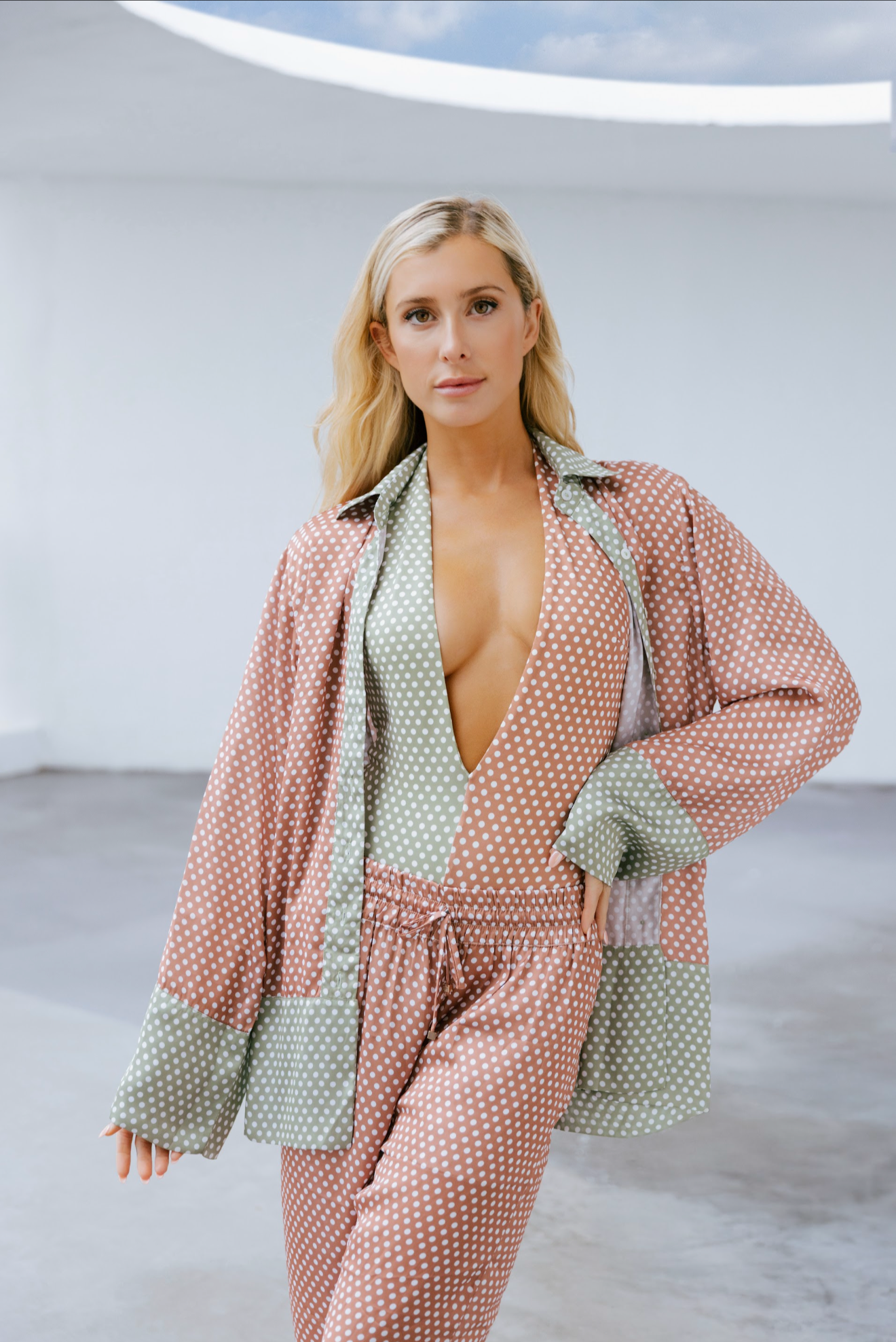 A woman with blonde hair wearing a pink and green polka dot pajama set in an indoor studio setting.