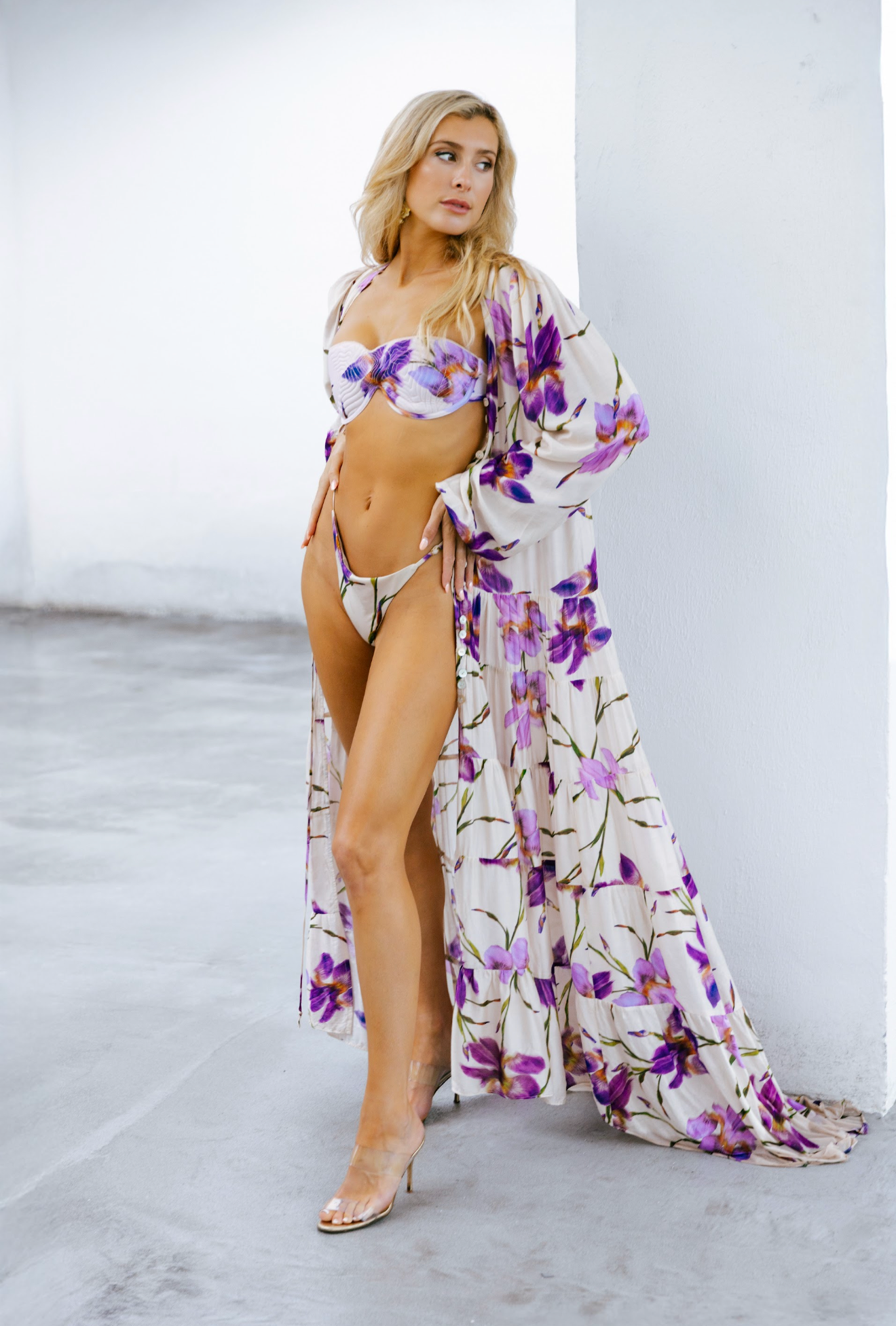 A woman with long blonde hair posing in a floral swimsuit with a matching cover-up against a white wall, wearing high heels.
