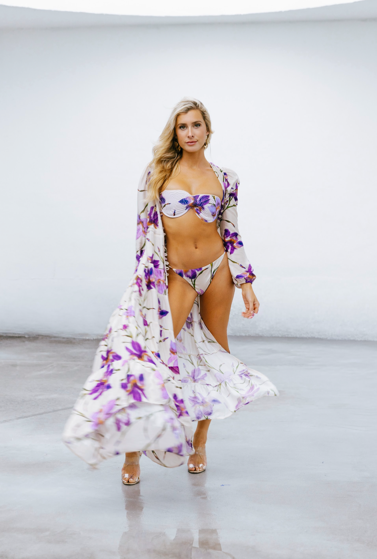 Allegra Paris A woman wearing a floral bikini and matching cover-up standing on a concrete floor in front of a white wall.