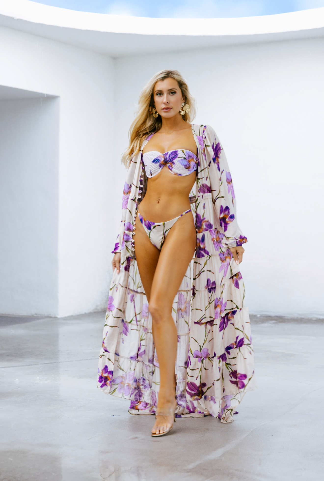A woman in a floral bikini and matching long cover-up walking towards the camera in a minimalist white space with a curved ceiling.