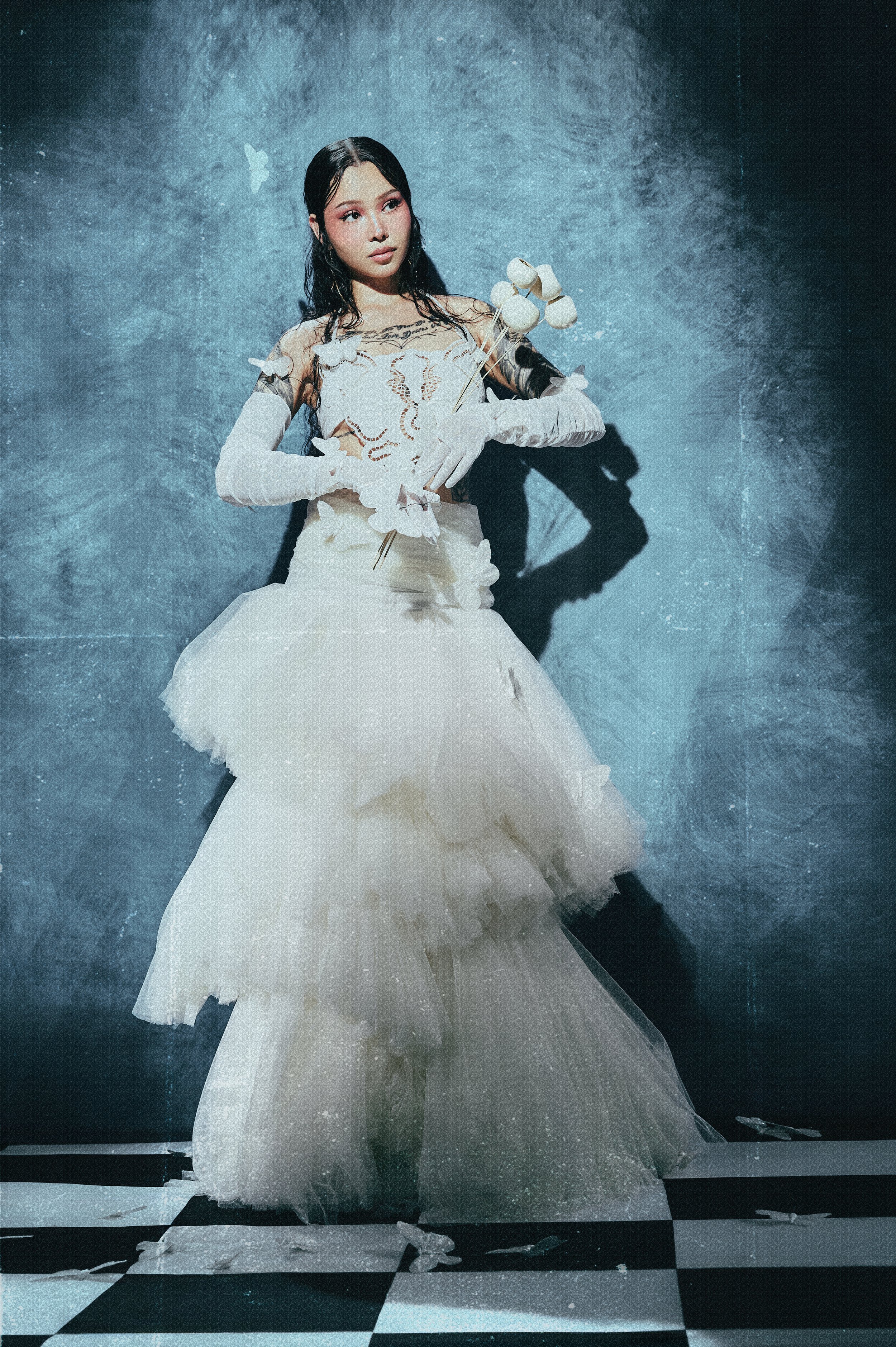 A woman in a white, ruffled gown stands against a textured blue background with butterflies, holding a skewer with marshmallows, casting a shadow on the wall.