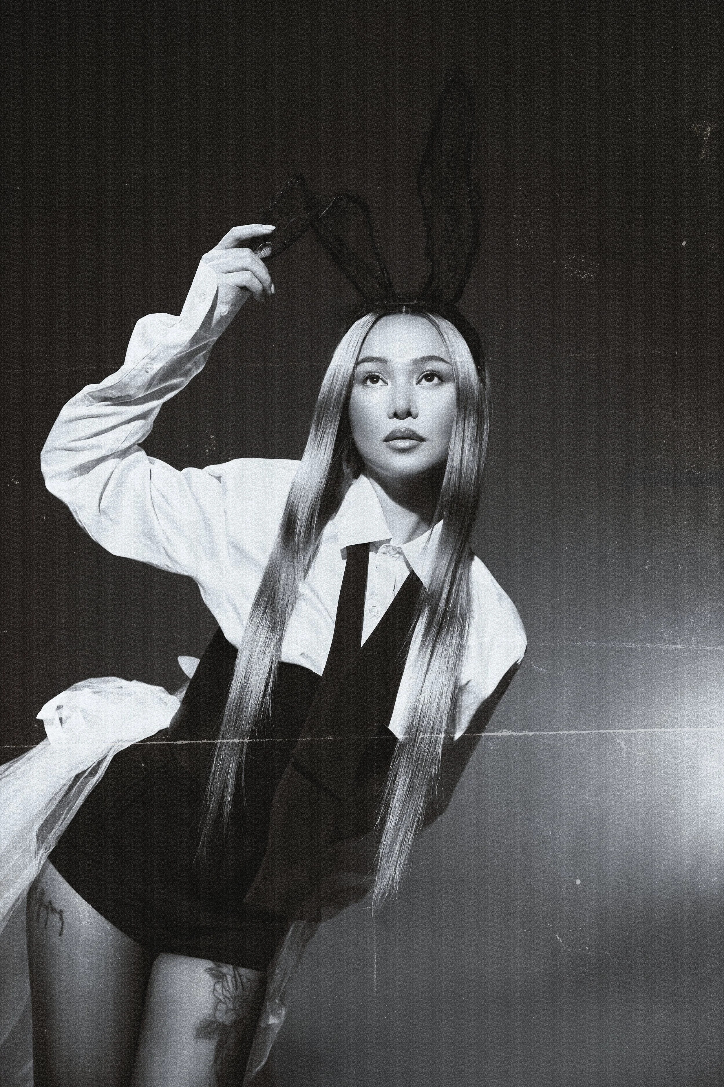 Black and white photo of a woman with long straight hair, wearing a white shirt and black shorts, holding bunny ears on her head, with tattoos visible on her thigh.