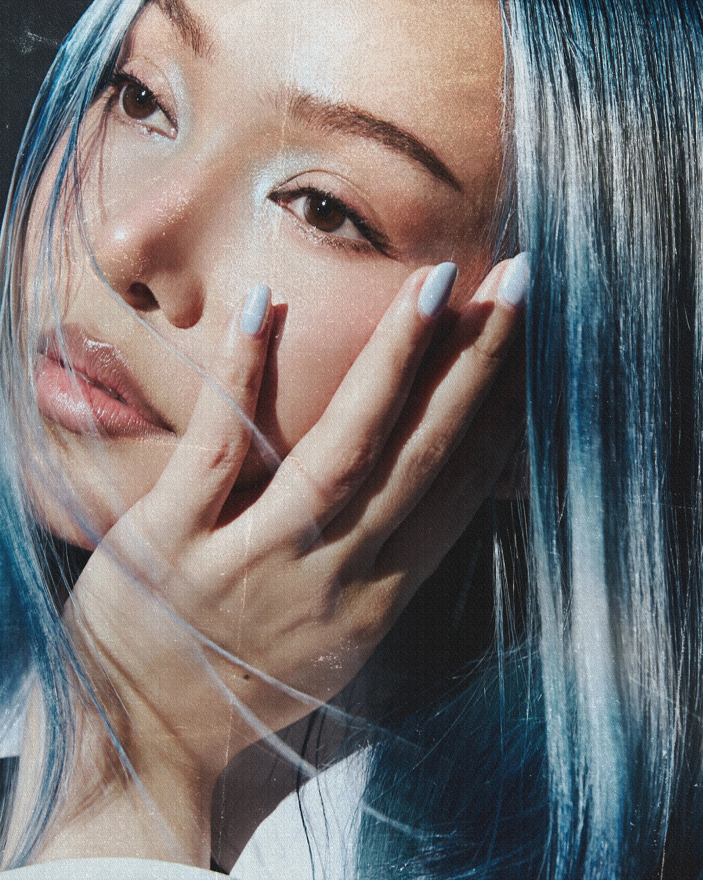 A close-up of a young woman with tan skin and long black and blue hair, touching her face with her hand, looking contemplative.