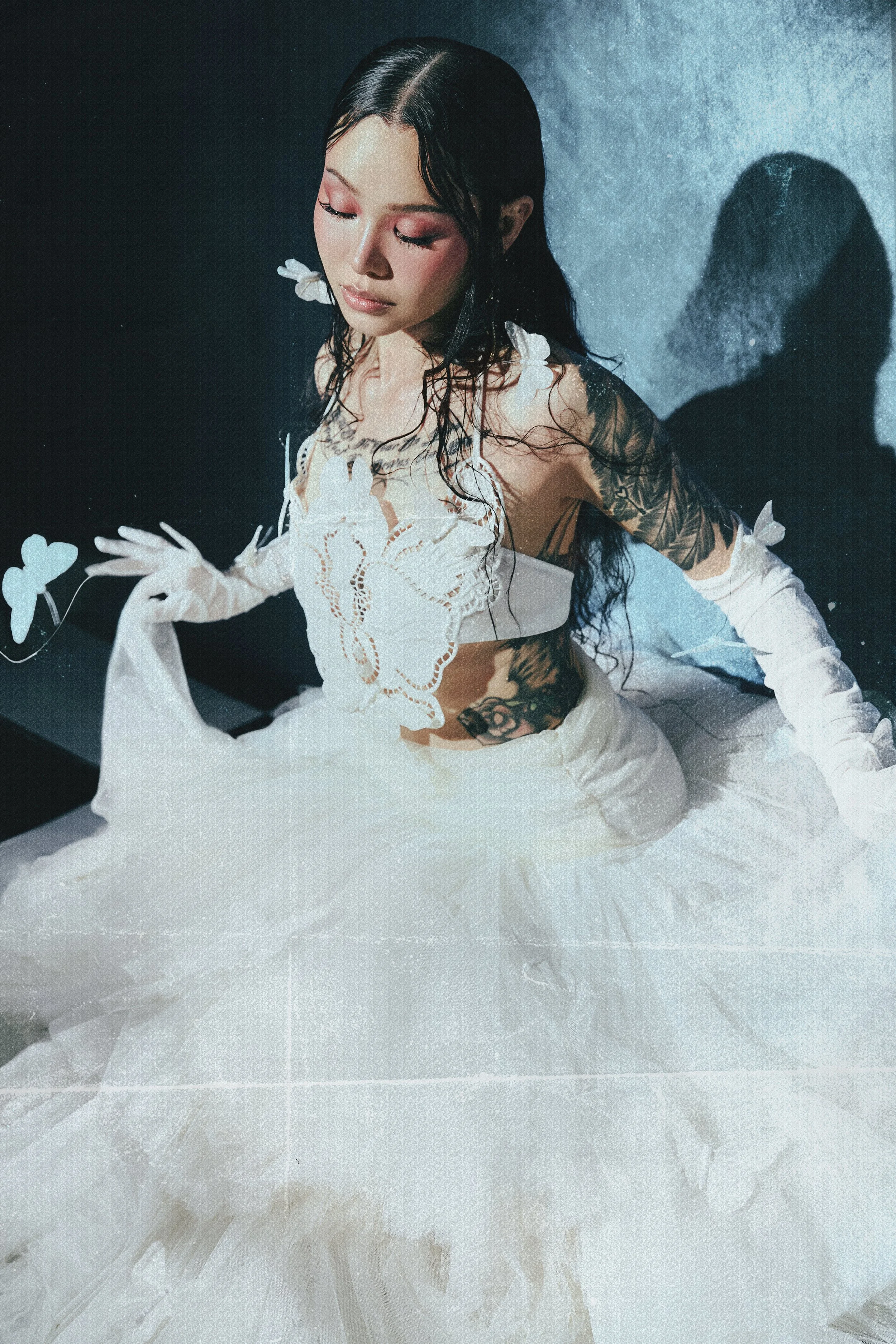 A woman with long wet black hair and closed eyes, wearing a white lace dress with butterfly details, is sitting on a textured surface. She has tattoos on her left arm and side. There are white butterfly decorations around her. Shadows and soft lighti
