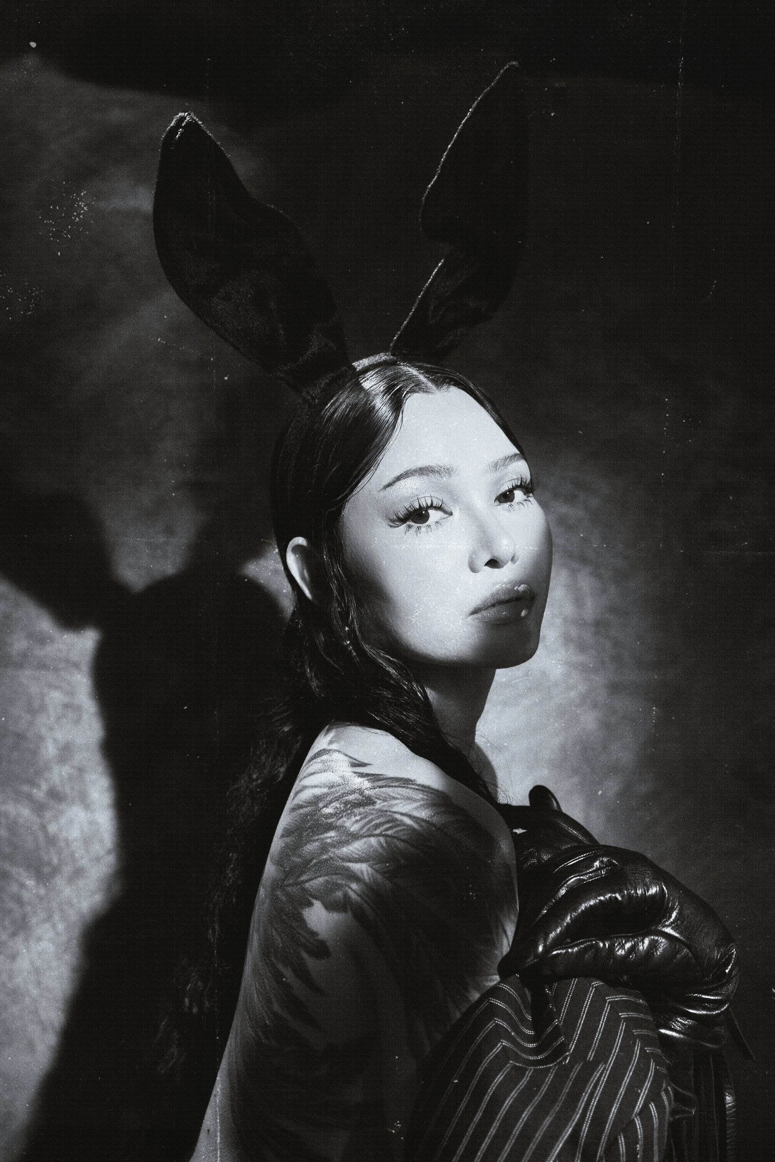 Black and white photo of a woman with long hair, wearing bunny ear headband, a dress with floral pattern, leather gloves, and striped pants, sitting on the floor with shadows cast on the background.