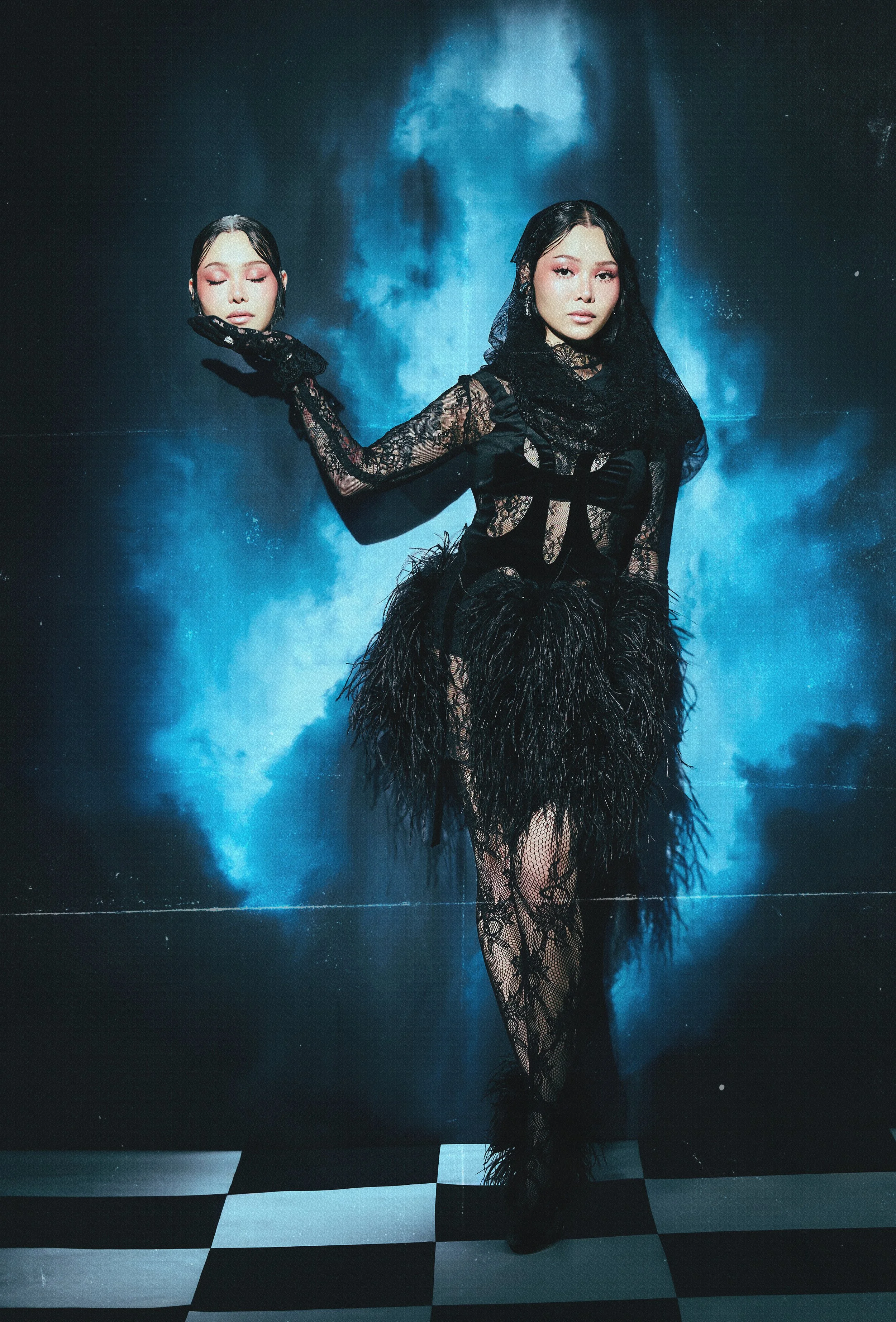 Woman dressed in black lace dress and feathered skirt, standing on black and white checkered floor, with blue smoke or clouds behind her.