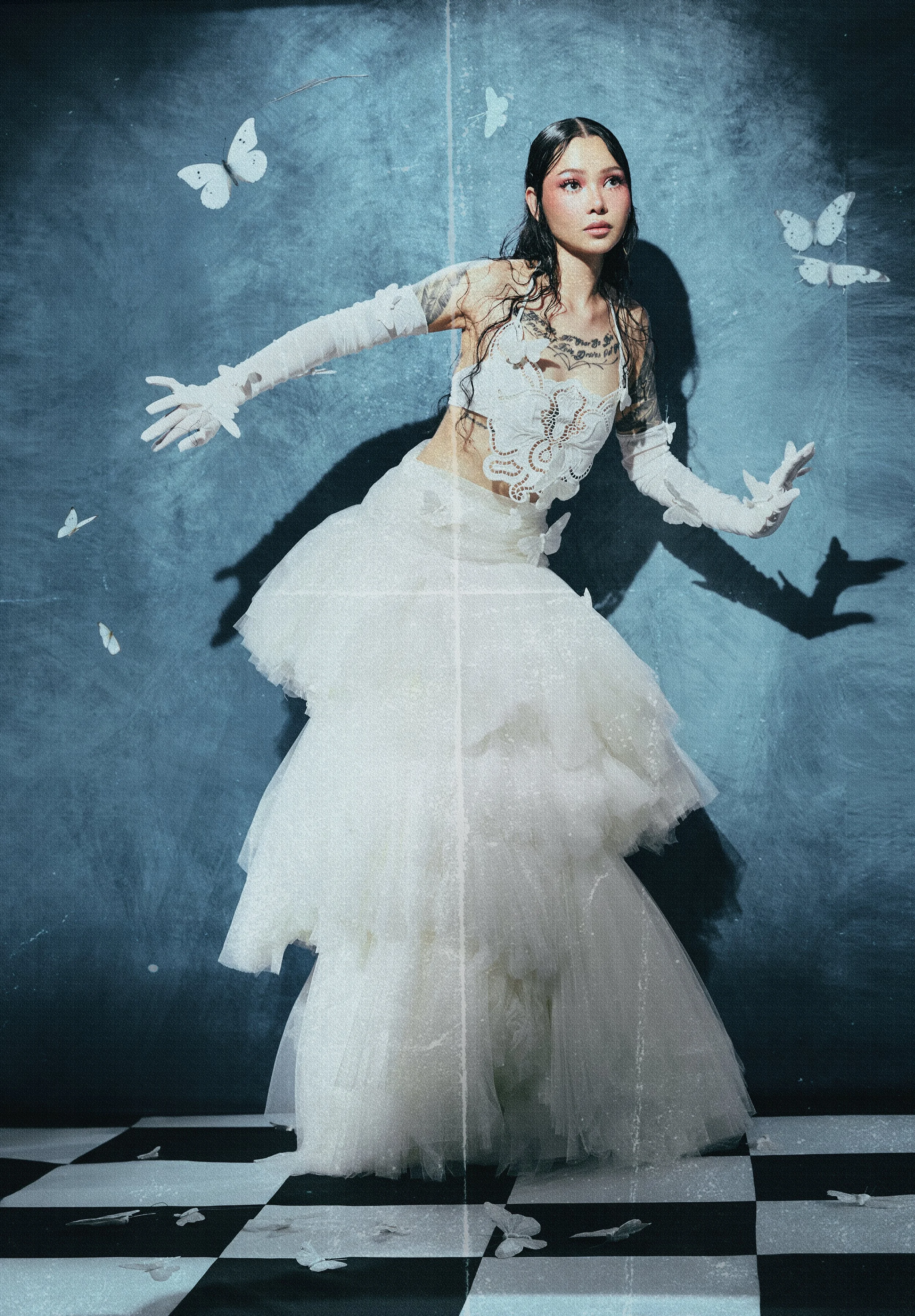 A woman in a white dress with lace and embroidery, long gloves, with butterflies in the background and on her dress, standing on a checkered floor with a textured blue wall behind her.
