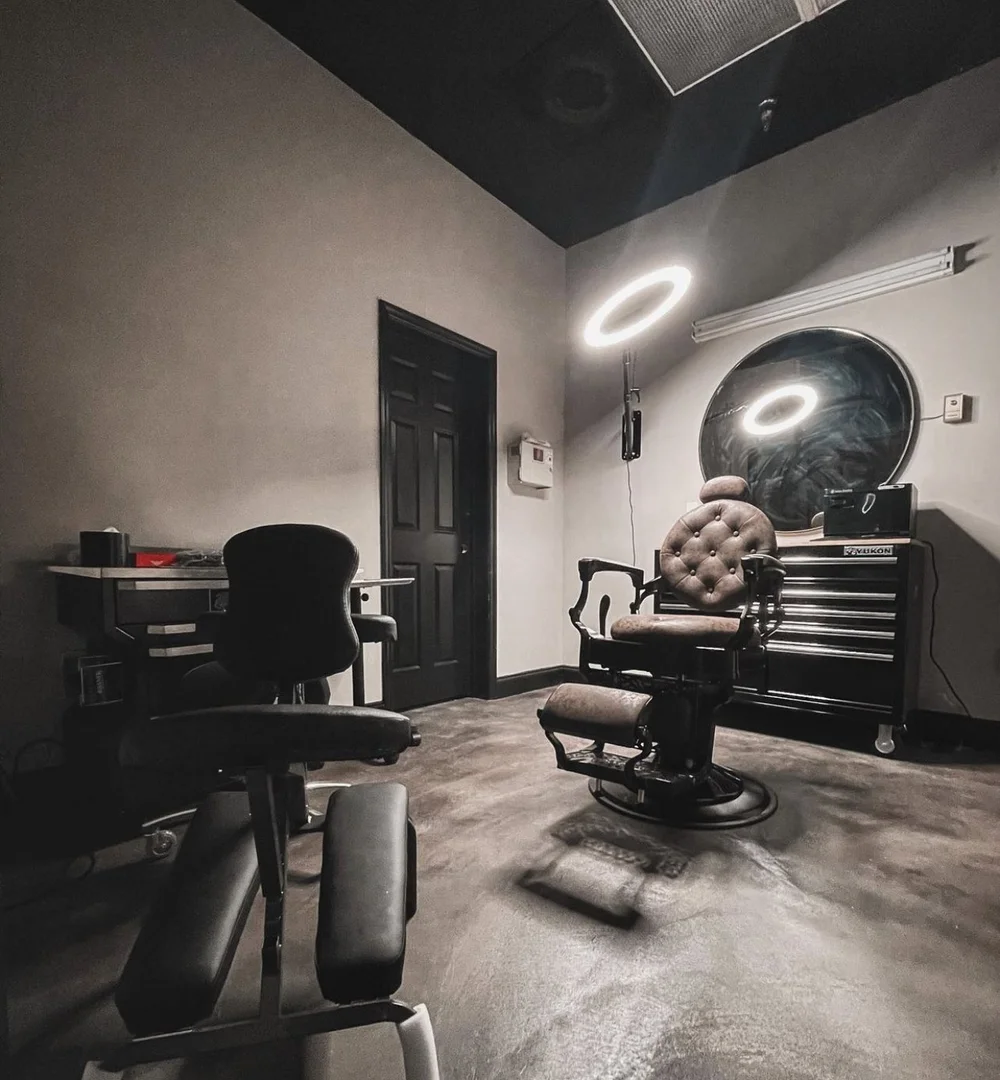 A treatment room with a reclining chair, a tufted leather barber chair, ring lights, a round mirror and a rolling tool chest.