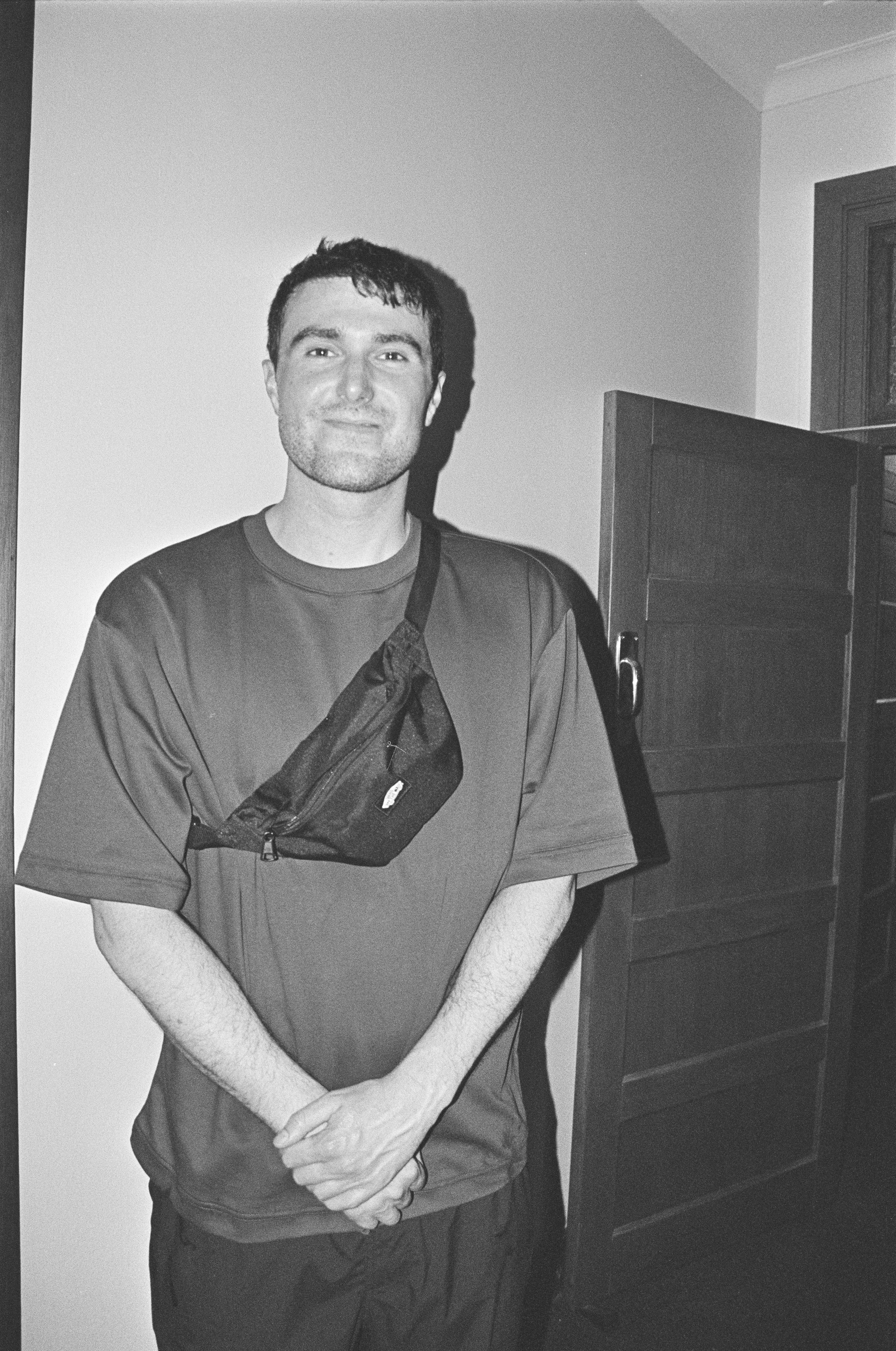 Black and white photo of a fred again standing in a room, wearing a T-shirt and a crossbody bag.