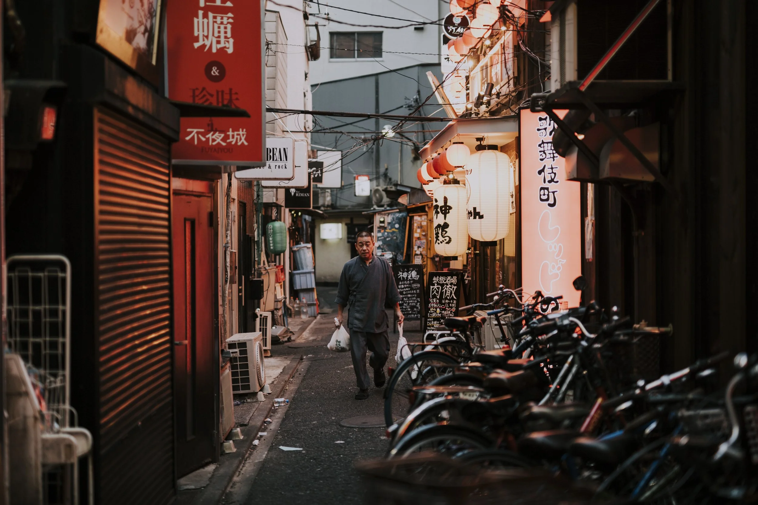 A narrow Japanese alley at night with shops and lanterns, featuring a person walking while carrying bags. There are several bicycles parked along the alleyway.