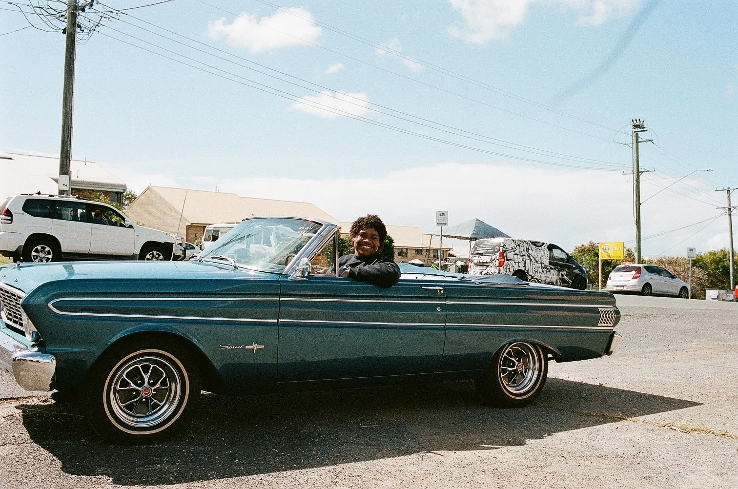 Person smiling while seated in a blue vintage convertible car parked on a sunny street, with other vehicles and buildings in the background.
