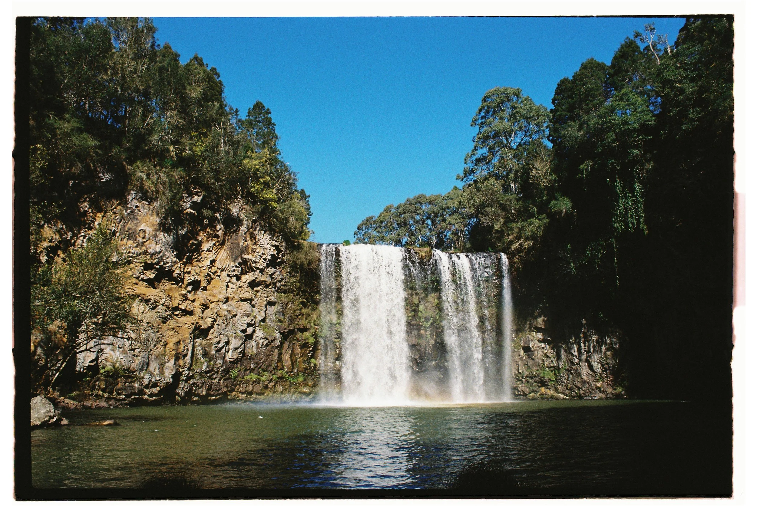 Waterfall flowing into a pool surrounded by lush green trees under a clear blue sky.