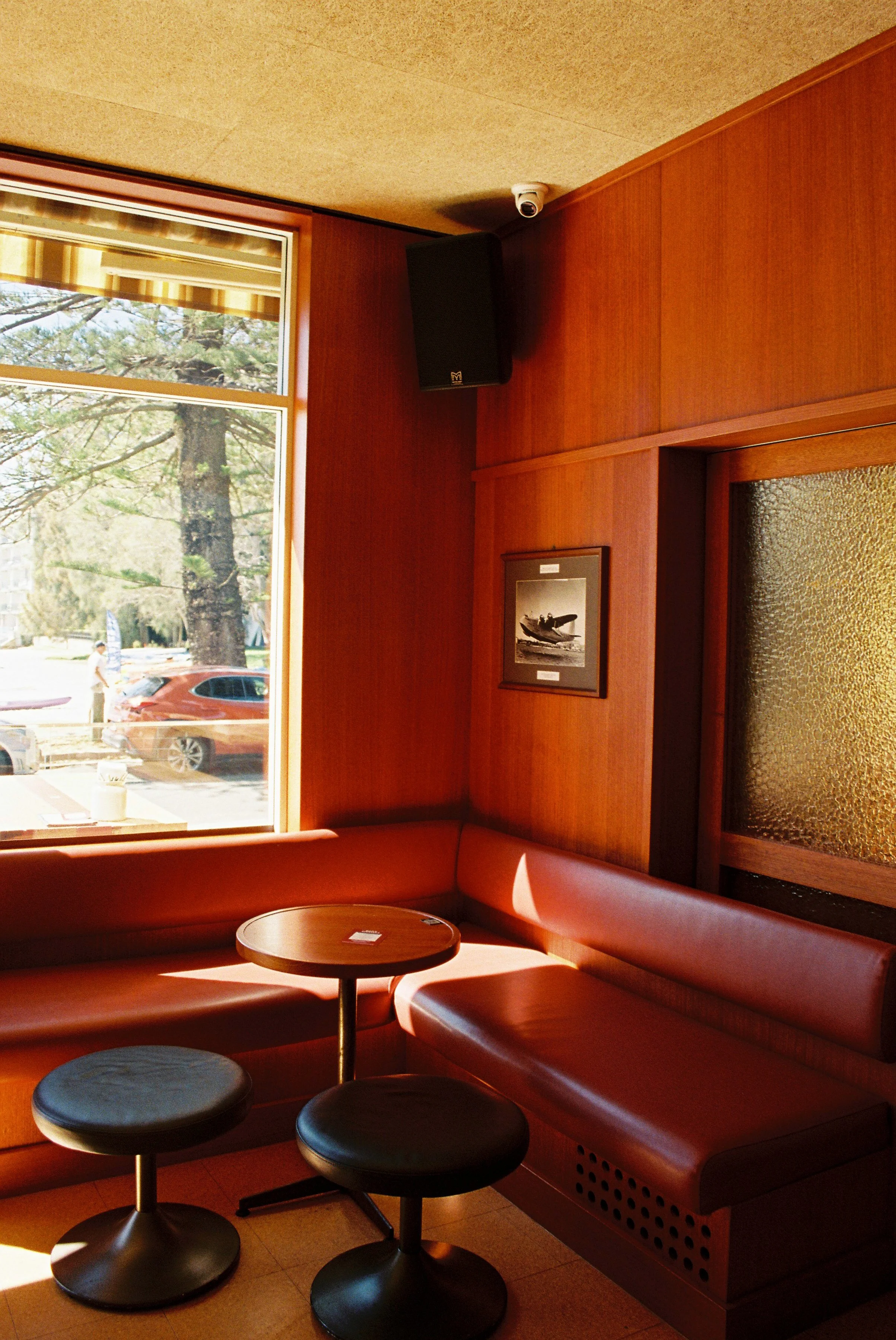 Interior of a café or diner with red booth seating, small circular tables with black stools, and a large window showing trees and cars outside. Club Rose Bay Photography