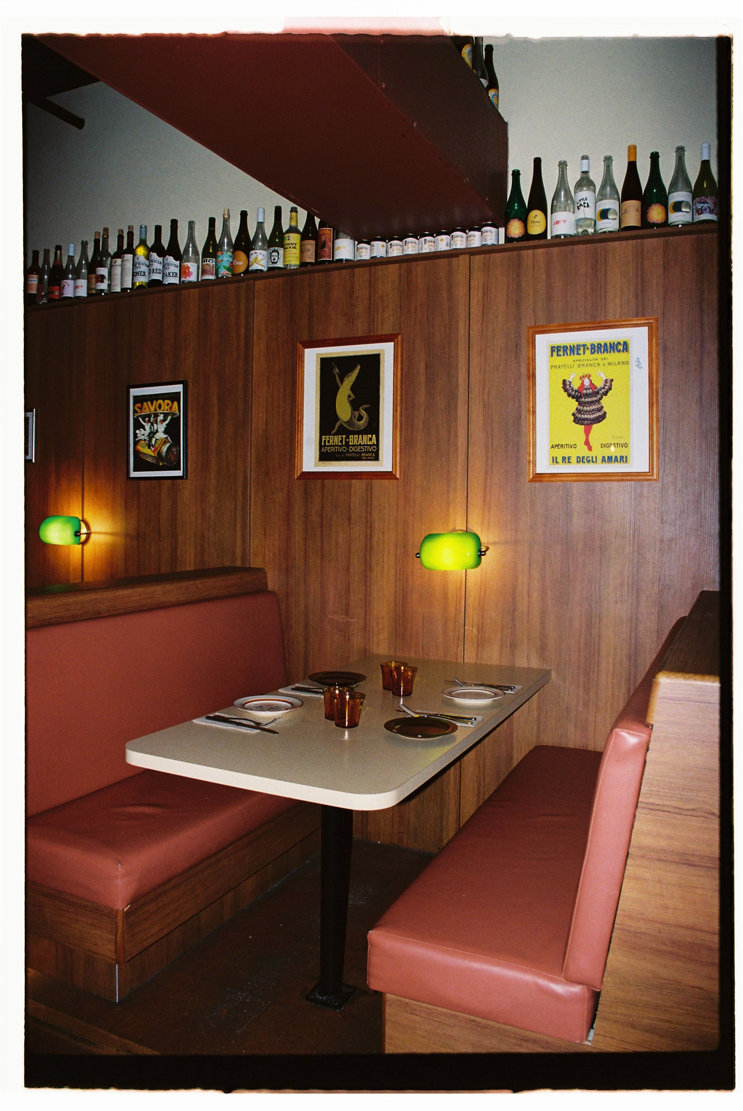 Retro diner booth with wood paneling, green wall lamps, and framed vintage posters. Table set with plates, cutlery, and glasses. Bottles displayed on a shelf above. Gold Coast | Byron Bay Photography by Joey Bailey