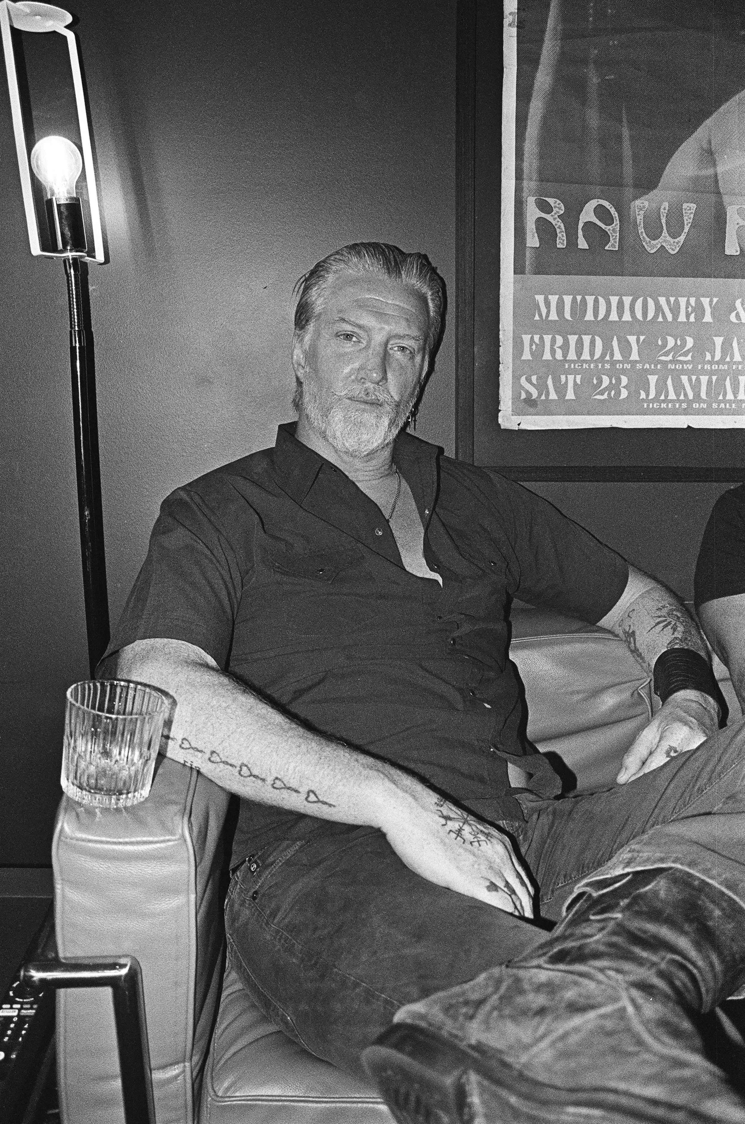Black and white photo of Josh Homme with tattoos sitting on a couch next to a drink and a lamp, with a poster on the wall behind him. Gold Coast | Byron Bay Photography by Joey Bailey