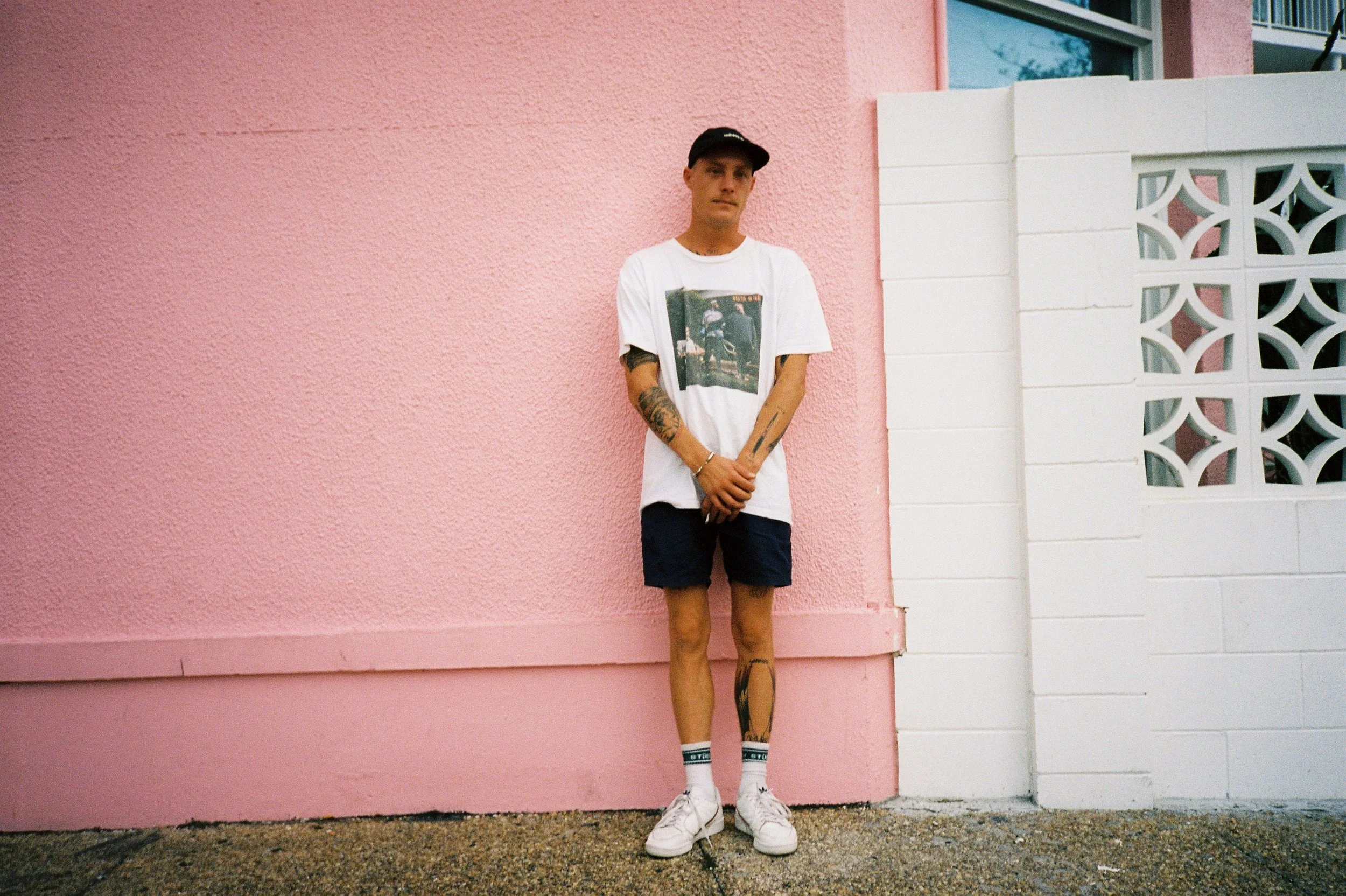 Person in casual attire standing against a pink wall, wearing a cap, graphic t-shirt, shorts, and sneakers, with tattoos visible on arms and legs.