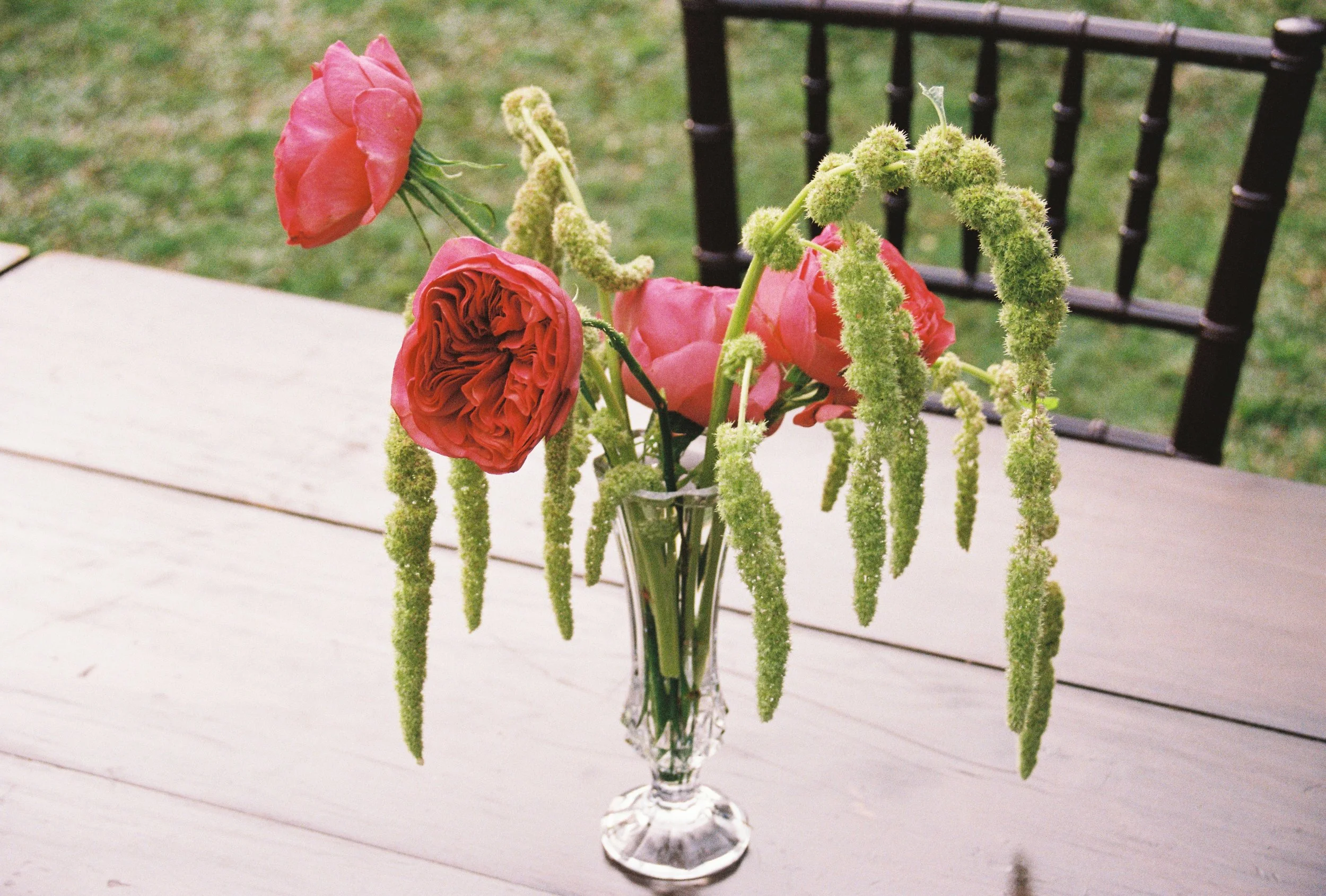 A bouquet of pink and coral roses and green veronica flowers in a crystal vase on a white table, with a green grassy background and black metal patio furniture.
