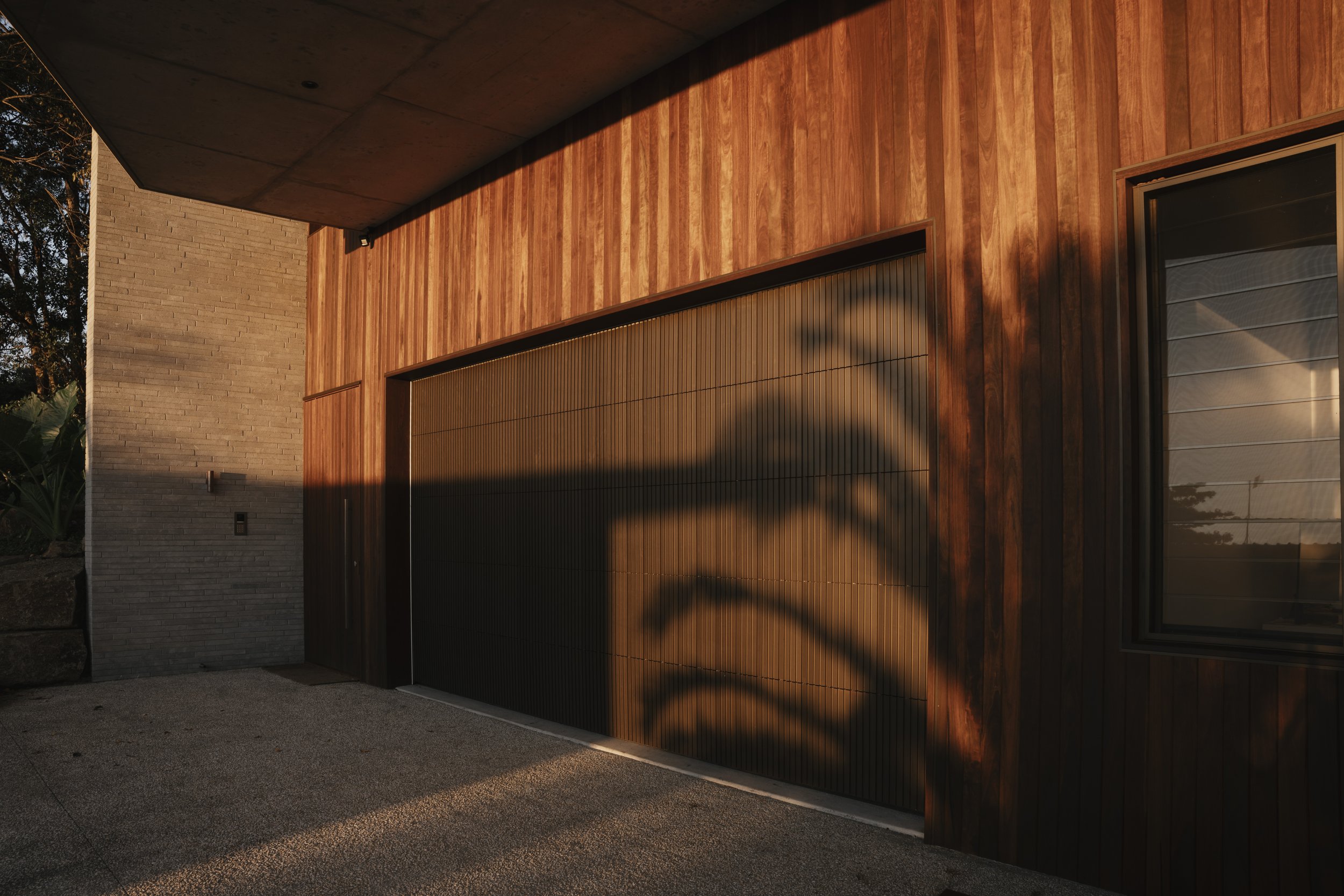 Modern wooden garage door with shadow of tree, concrete wall, and adjacent window in evening light.