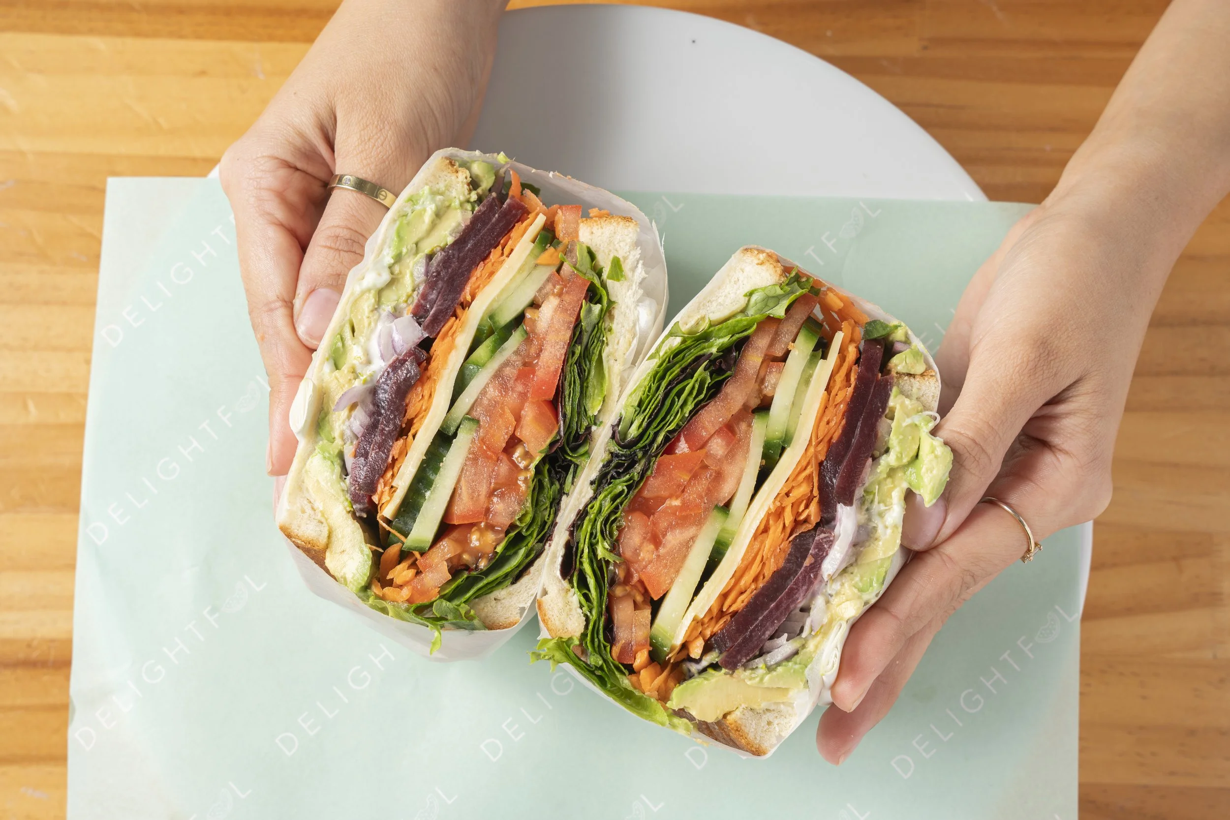 Two halves of a veggie sandwich with lettuce, tomato, cucumber, shredded carrots, and dark hummus in a bun, being held over a light blue delighful branded paper on a wooden surface. Gold Coast Food photography by Joey Bailey