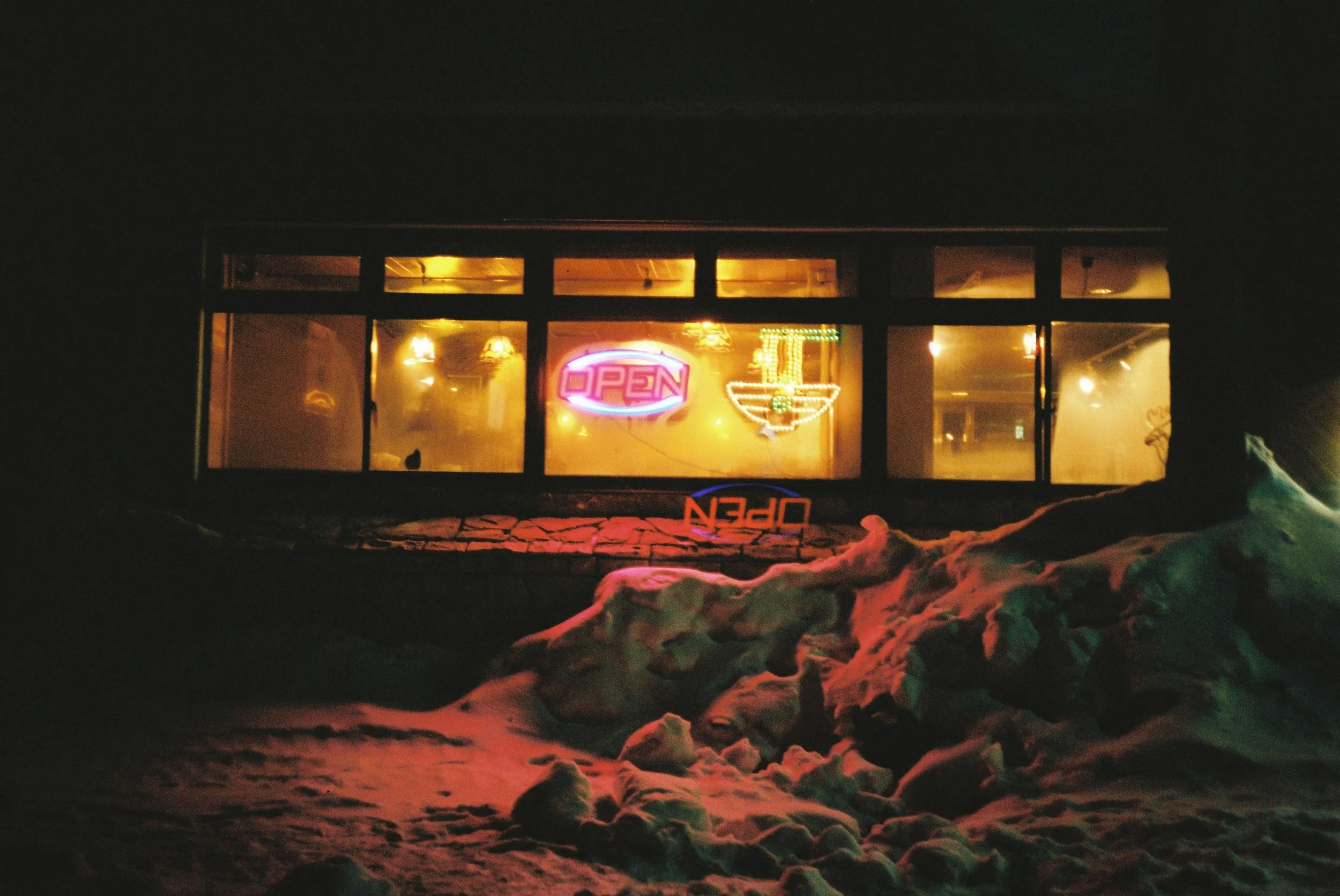 Snow-covered landscape in front of a building with neon lights and 'OPEN' sign at night.