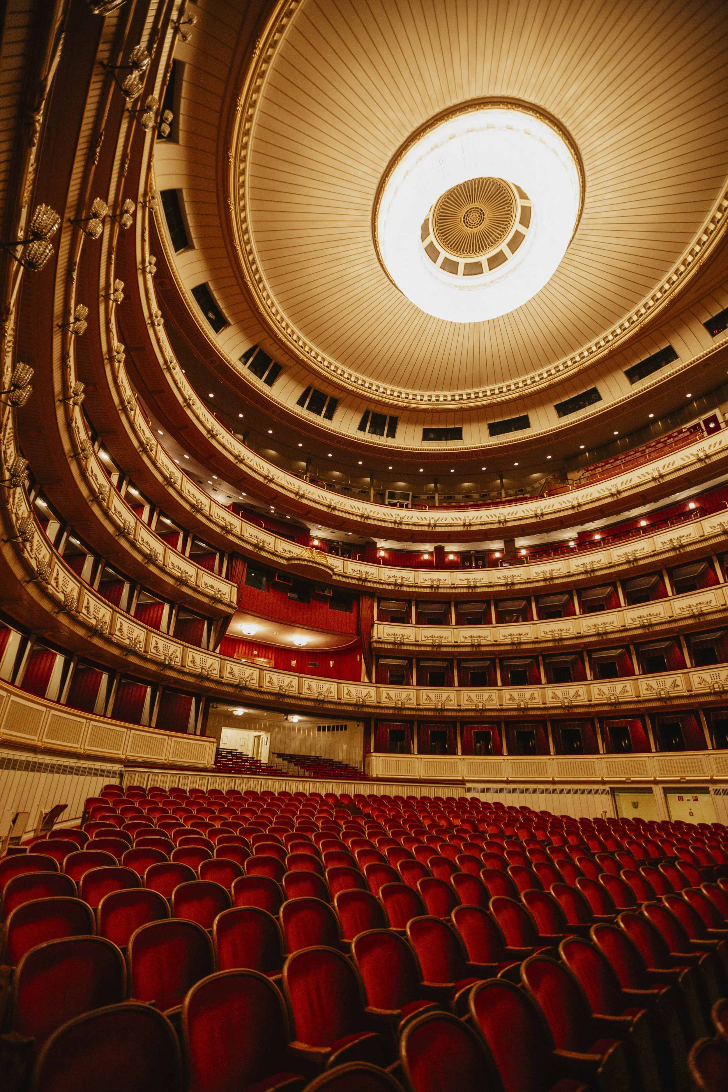 Interior of a grand theater with rows of red velvet seats and ornate, multi-tiered balconies extending toward a domed ceiling and chandelier.