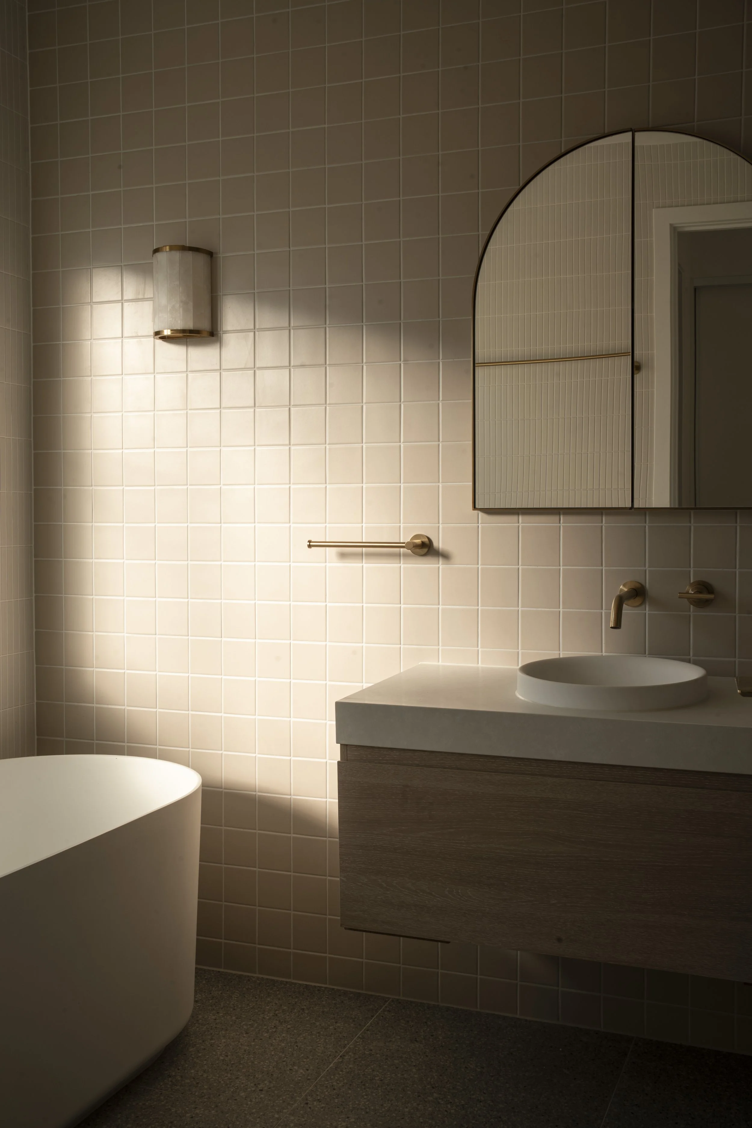 Modern bathroom with beige tiled walls, a white freestanding bathtub, a wooden vanity with a round vessel sink, gold fixtures, a wall-mounted cabinet with mirror, and a small wall sconce. Architectural photography by Joey Bailey, Gold Coast