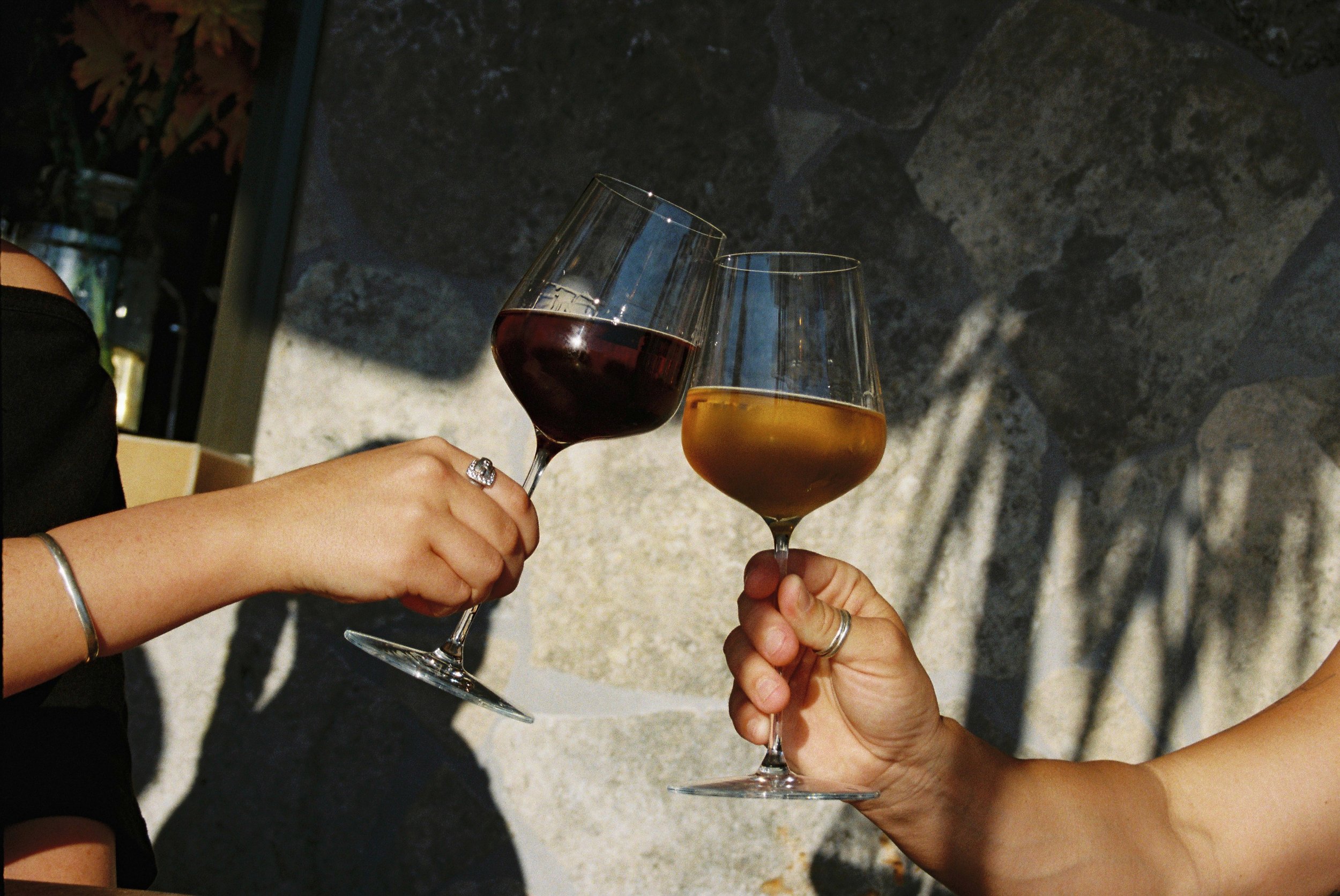 Two people clinking glasses of wine, one with red wine and the other with white wine, against a stone wall background. moustache mermaid beach food photography