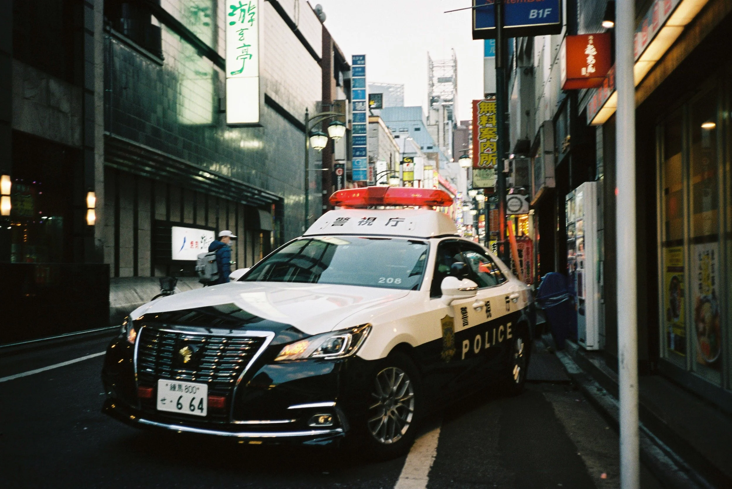 Japanese police car parked on a narrow urban street with illuminated signs and storefronts.