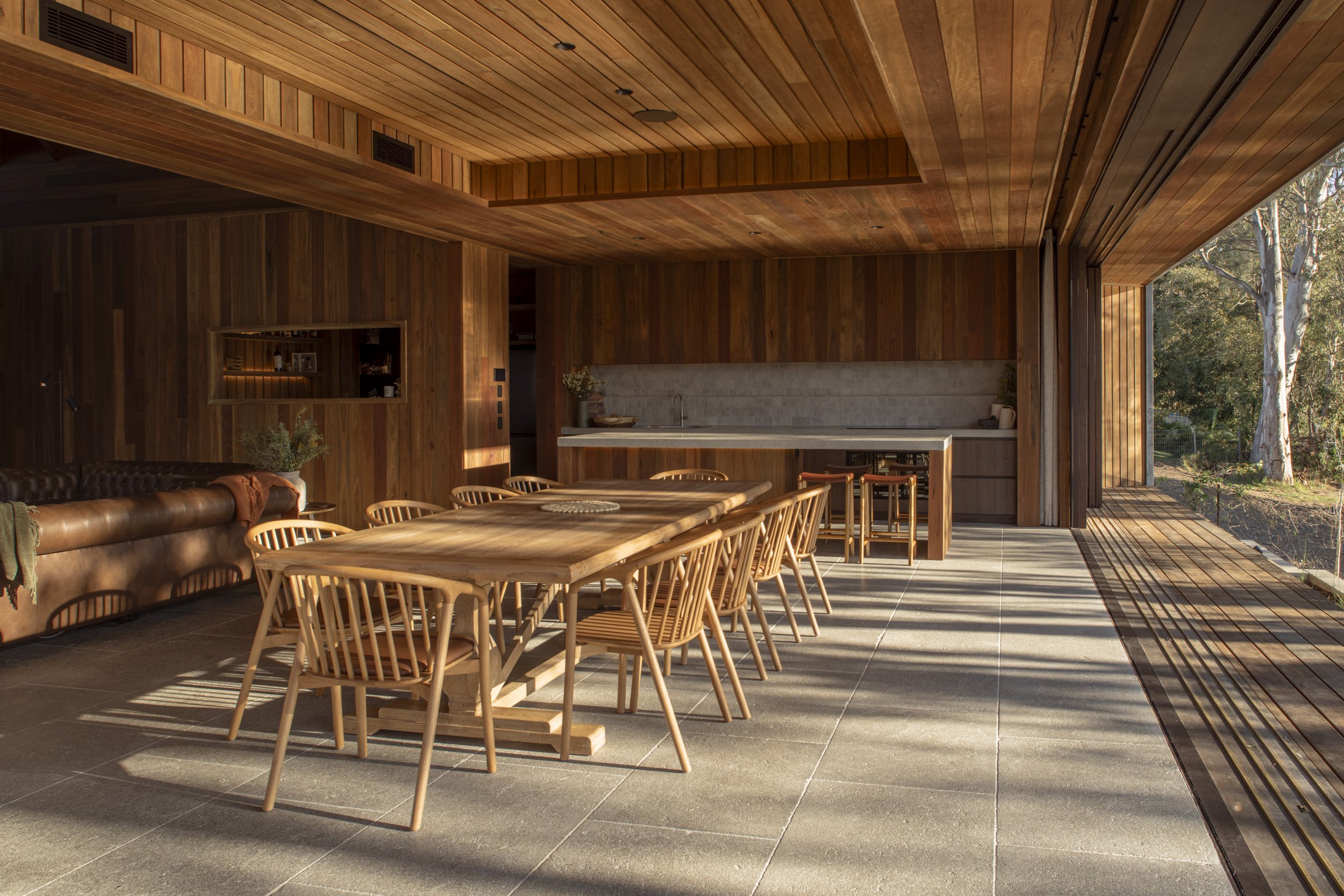 Modern dining area with wooden table and chairs, open kitchen with concrete countertop, and large glass wall overlooking outdoor trees. Gold Coast Architectural Photography by Joey Bailey 