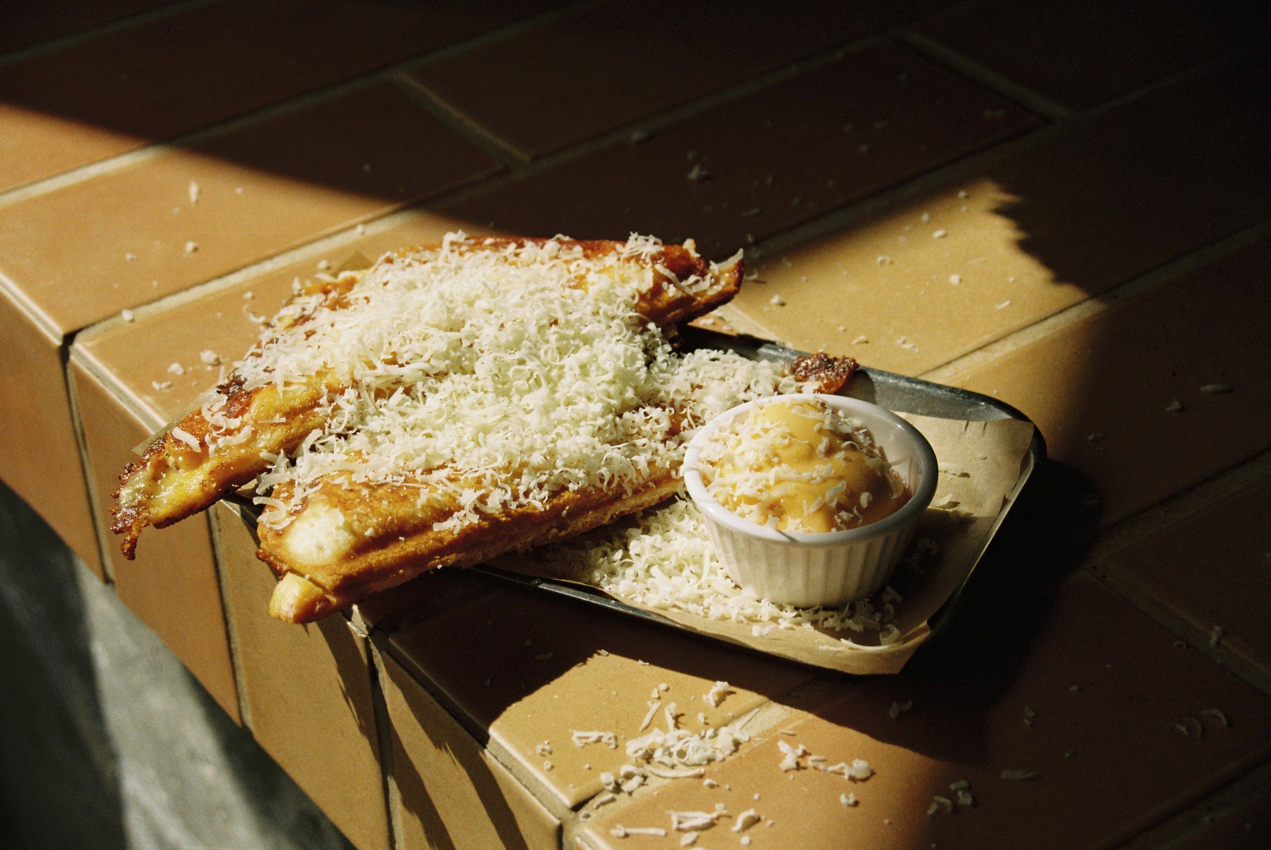 Cheese pizza slice with grated cheese, served with a side of cheese topping in a small bowl, on a cardboard tray.
