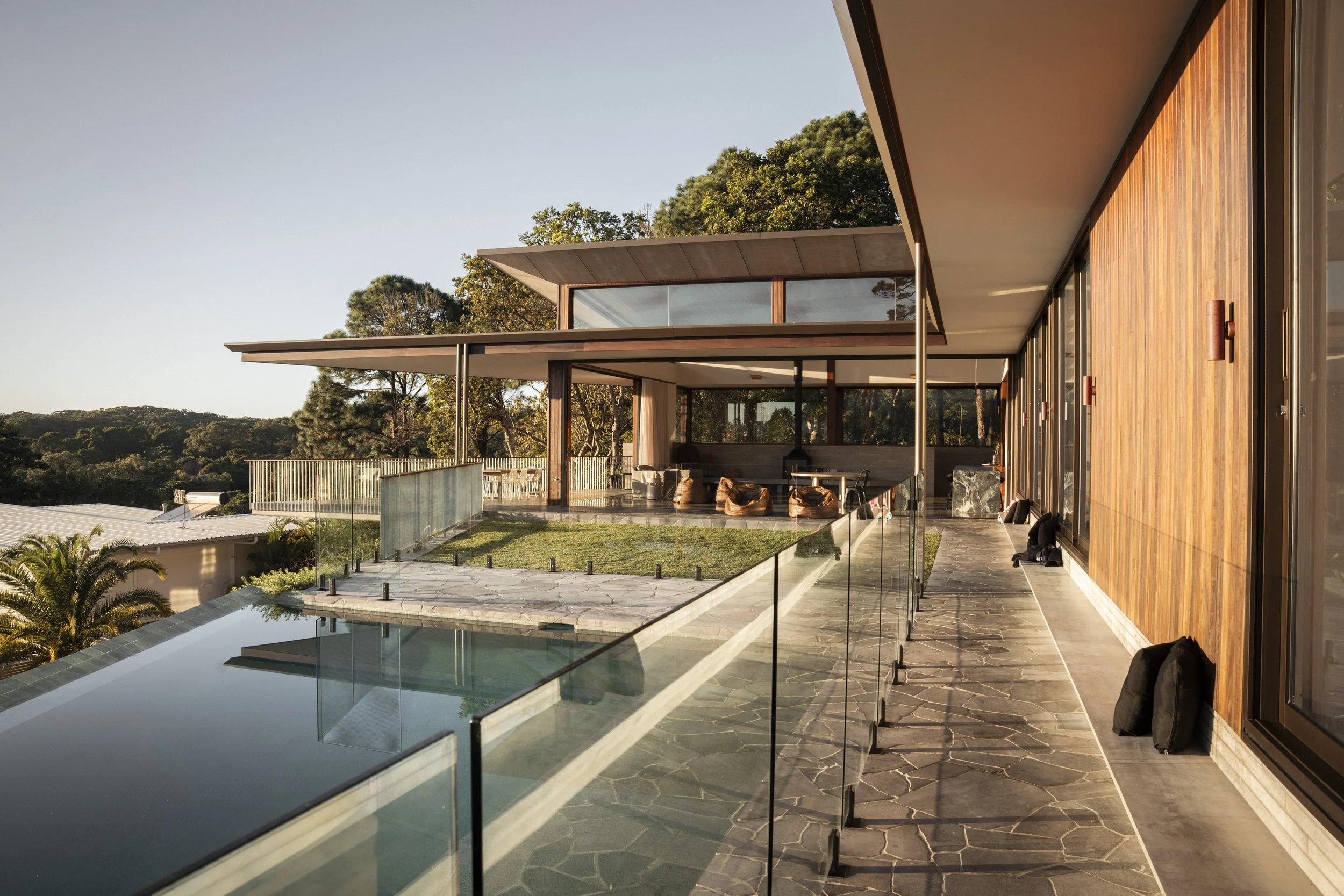 Modern house with a spacious outdoor balcony overlooking a lush green landscape, featuring glass railings, a swimming pool, and wood-paneled walls. Cape House, Suffolk Park – architectural photography