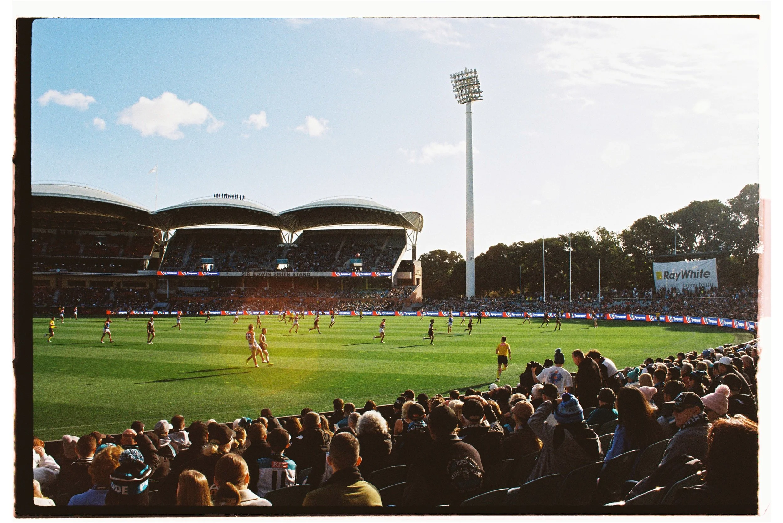 An Australian rules football game being played in a large stadium with spectators in the stands, a green field, and players in yellow and maroon uniforms.