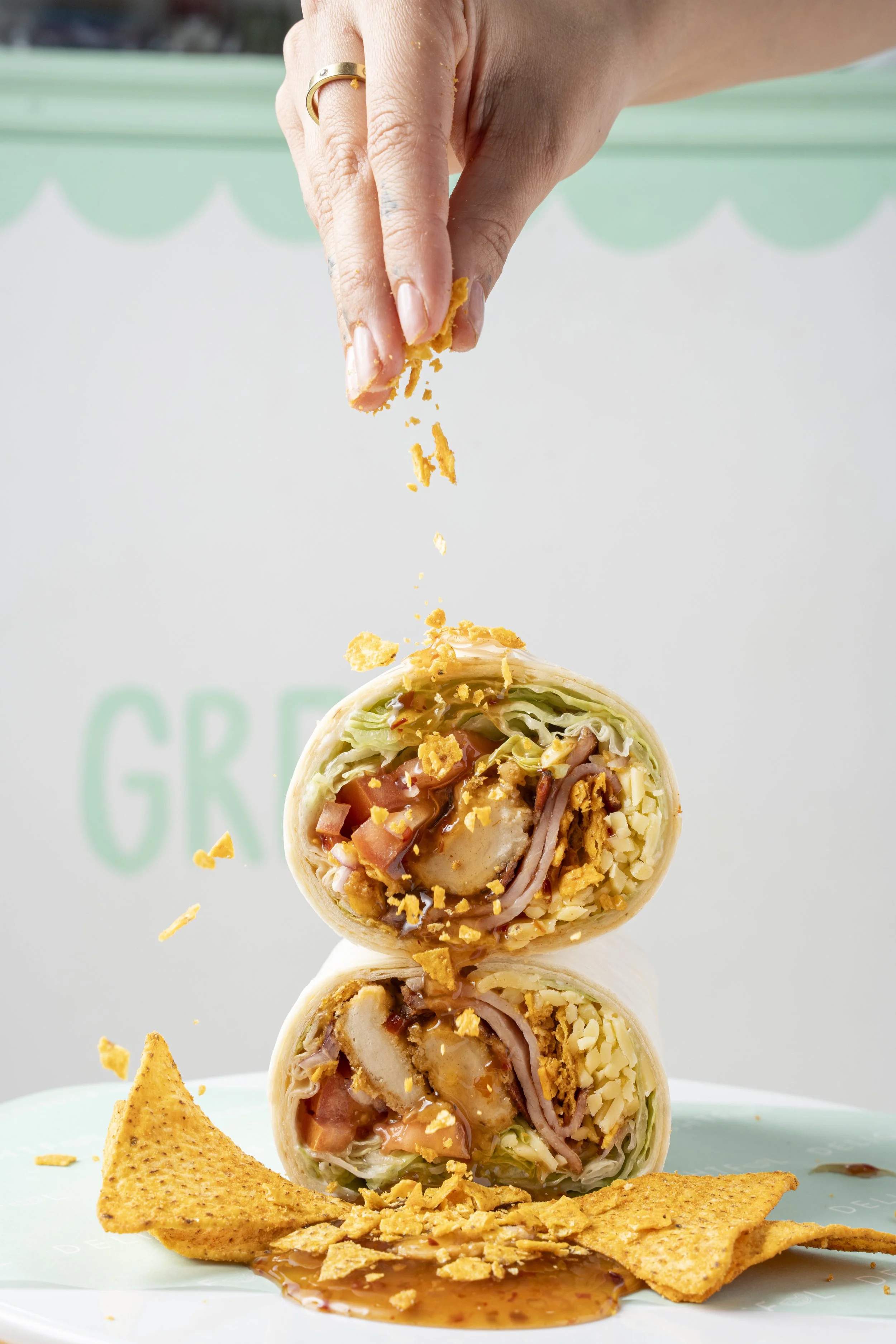 A hand sprinkling crushed tortilla chips over a stacked wrap cut in half, revealing chicken, bacon, lettuce, tomatoes, shredded cheese, and sauce inside, with tortilla chips and sauce on a plate below. Gold Coast Food photography by Joey Bailey
