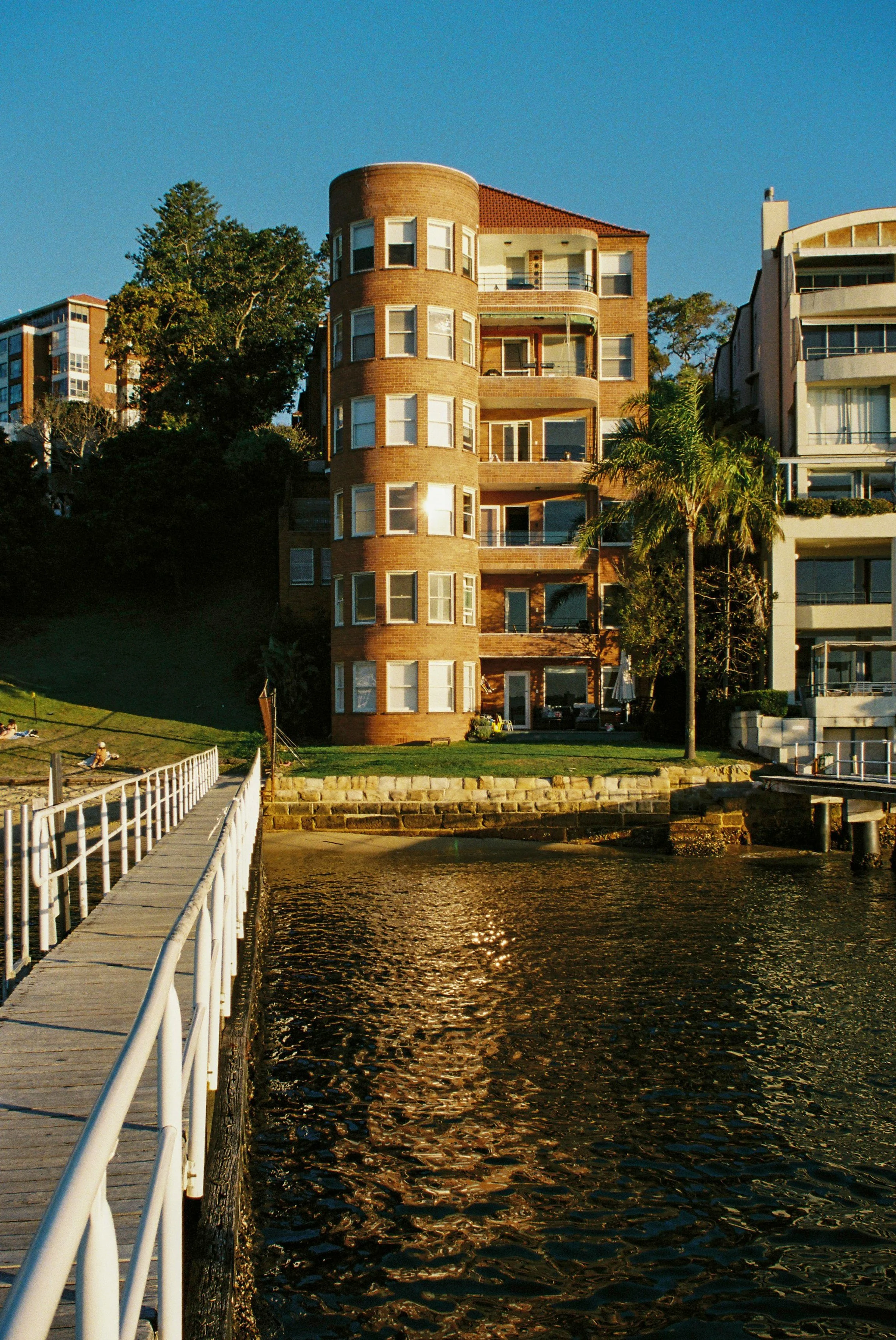 A waterfront scene featuring a wooden dock with white railings extending over dark water. In the background, there are multi-story residential buildings,Garron Tower Art Deco architecture Sydney Nsw