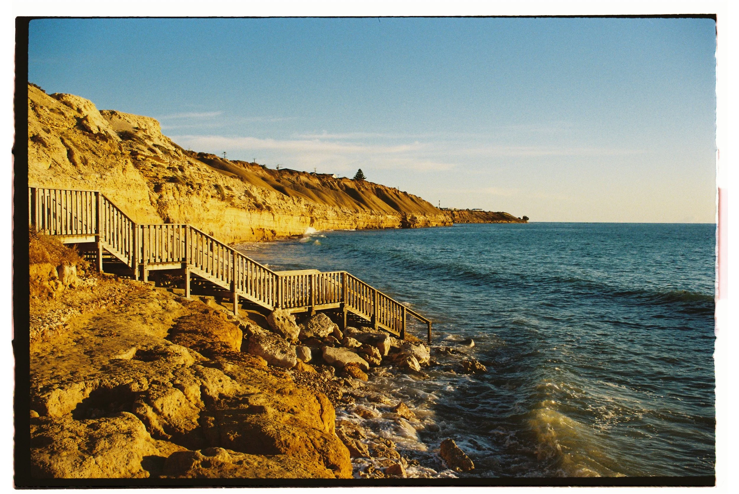 35mm photoWooden stairs leading down to a rocky shoreline along a sandy cliffside, with the ocean and a clear sky in the background.Gold Coast | Byron Bay Photography by Joey Bailey