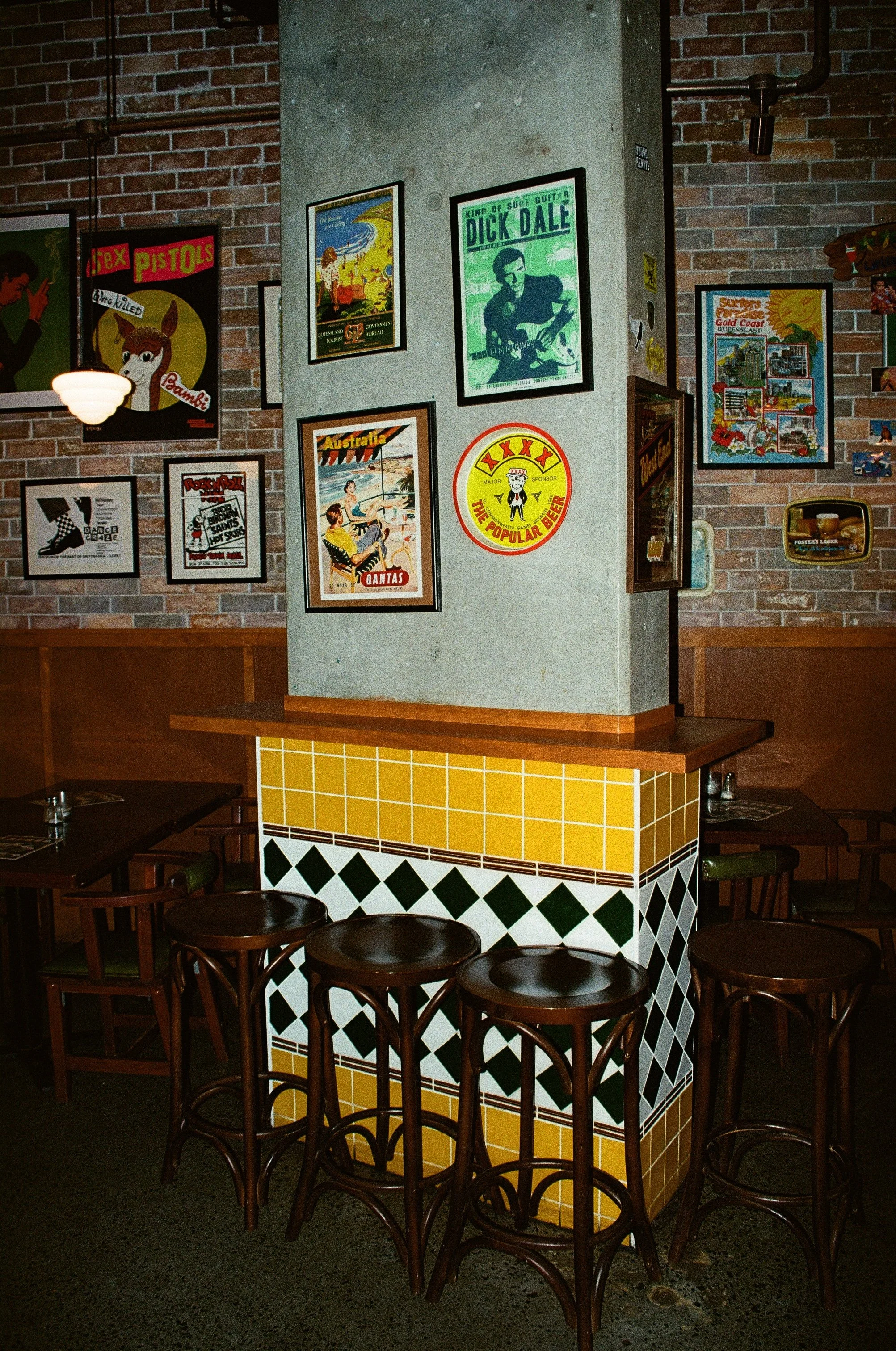 Interior of a restaurant or pub with a brick wall decorated with vintage posters and signs, including a picture of a guitar legend, a beach scene, and beer advertisements. There are wooden tables and stools, with a tiled column in the center.