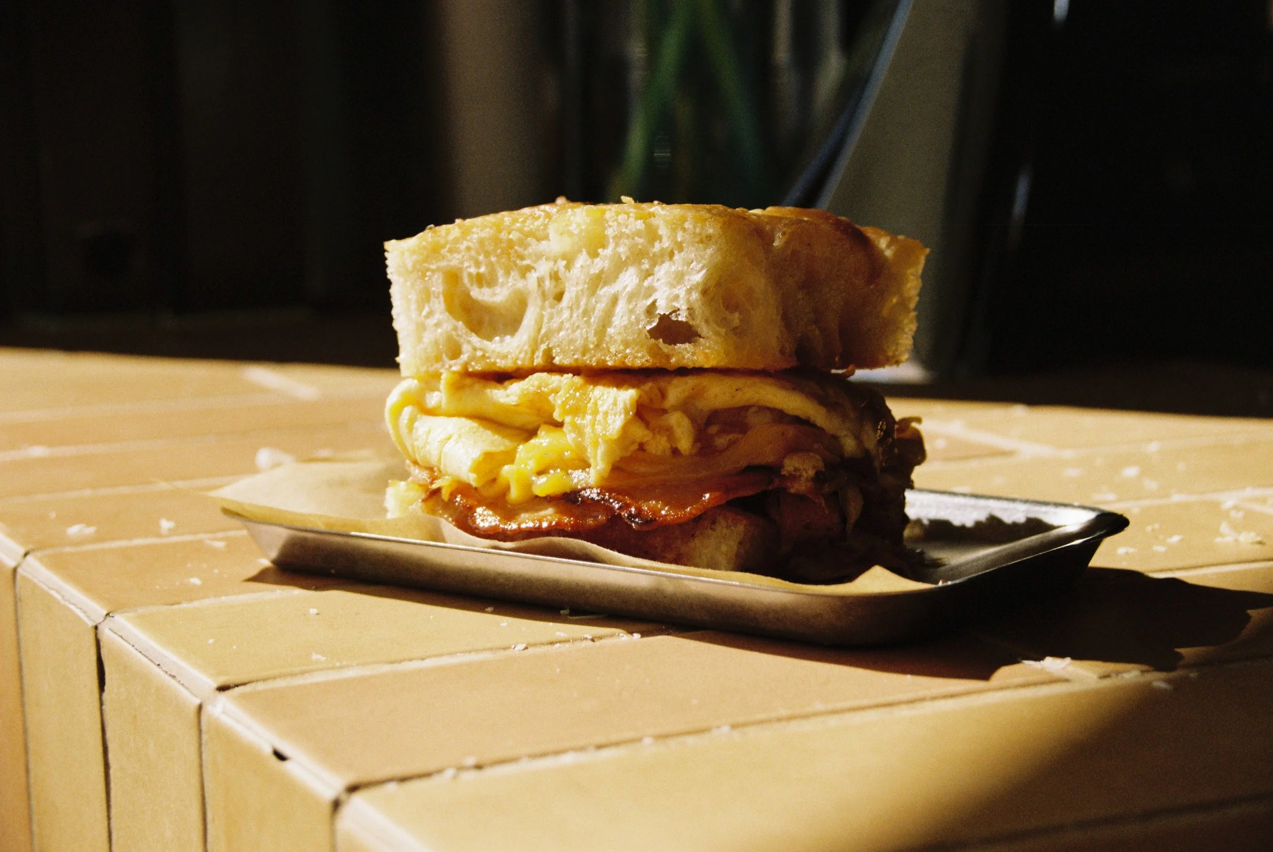 A sandwich with a slice of bread on top, containing eggs and bacon, on a plate placed on a wooden surface.