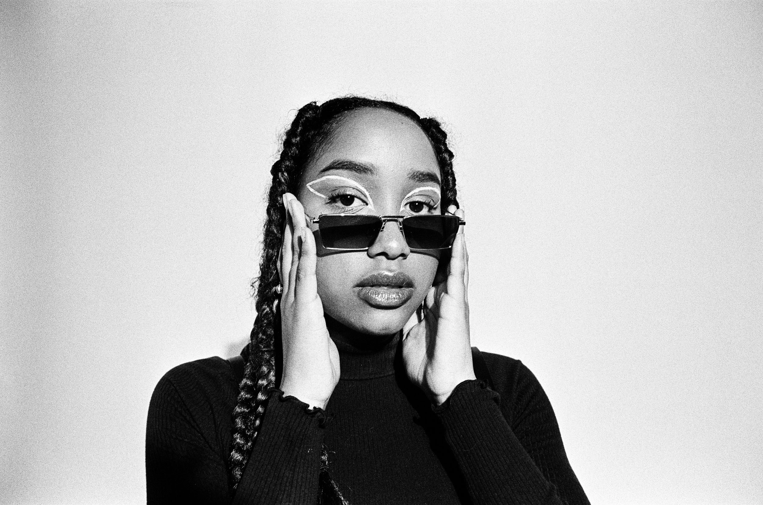 Black and white photo of a person wearing sunglasses and a black turtleneck, with hands near their face.