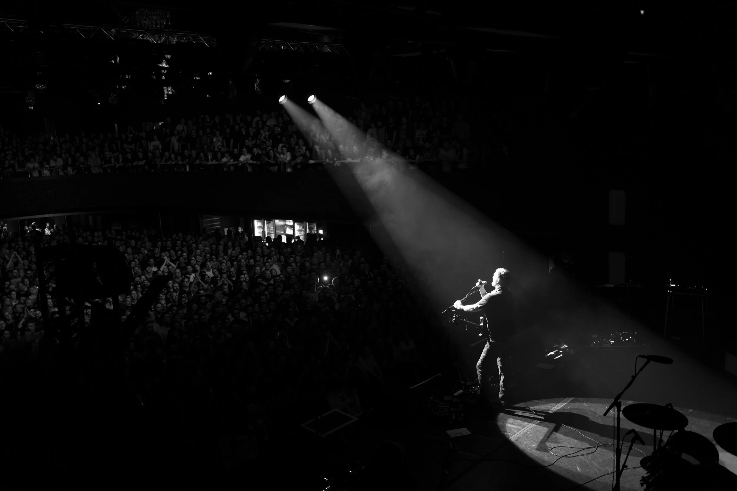 Black and white photo of a musician on stage with a spotlight, in front of a large audience in a concert venue.